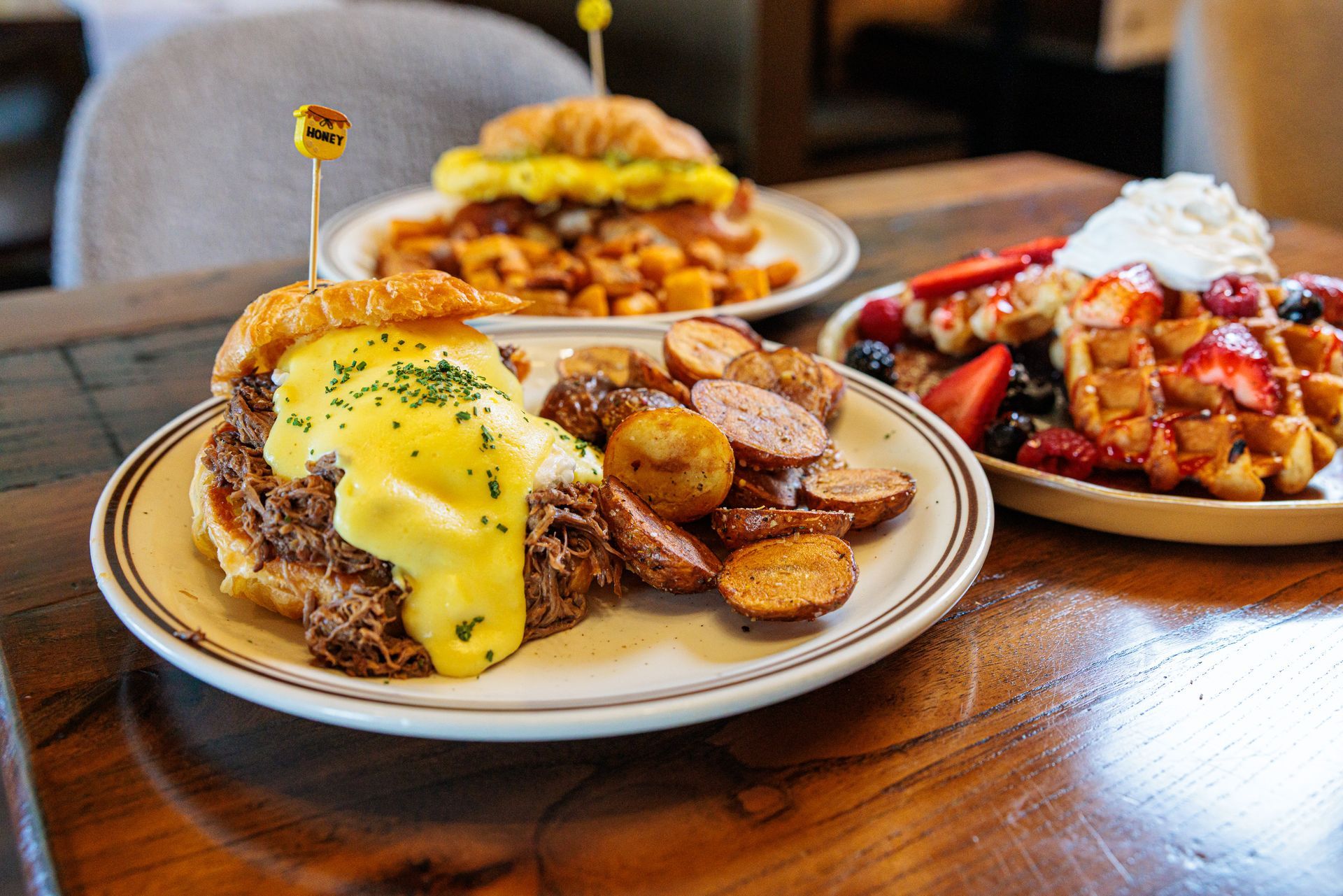 Plates of breakfast food on a wooden table: egg sandwiches, potatoes, and waffles topped with fruit and whipped cream.