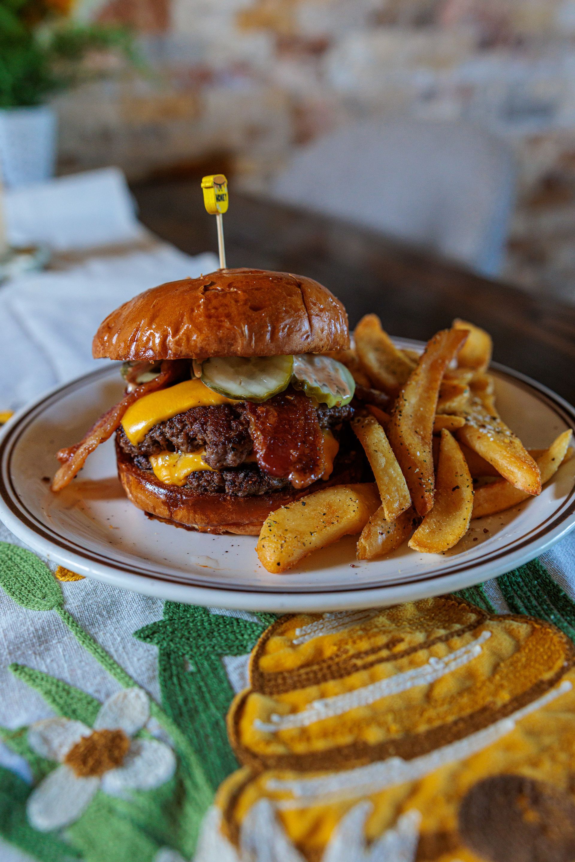 Double cheeseburger with bacon and pickles on a plate with fries. Yellow toothpick.