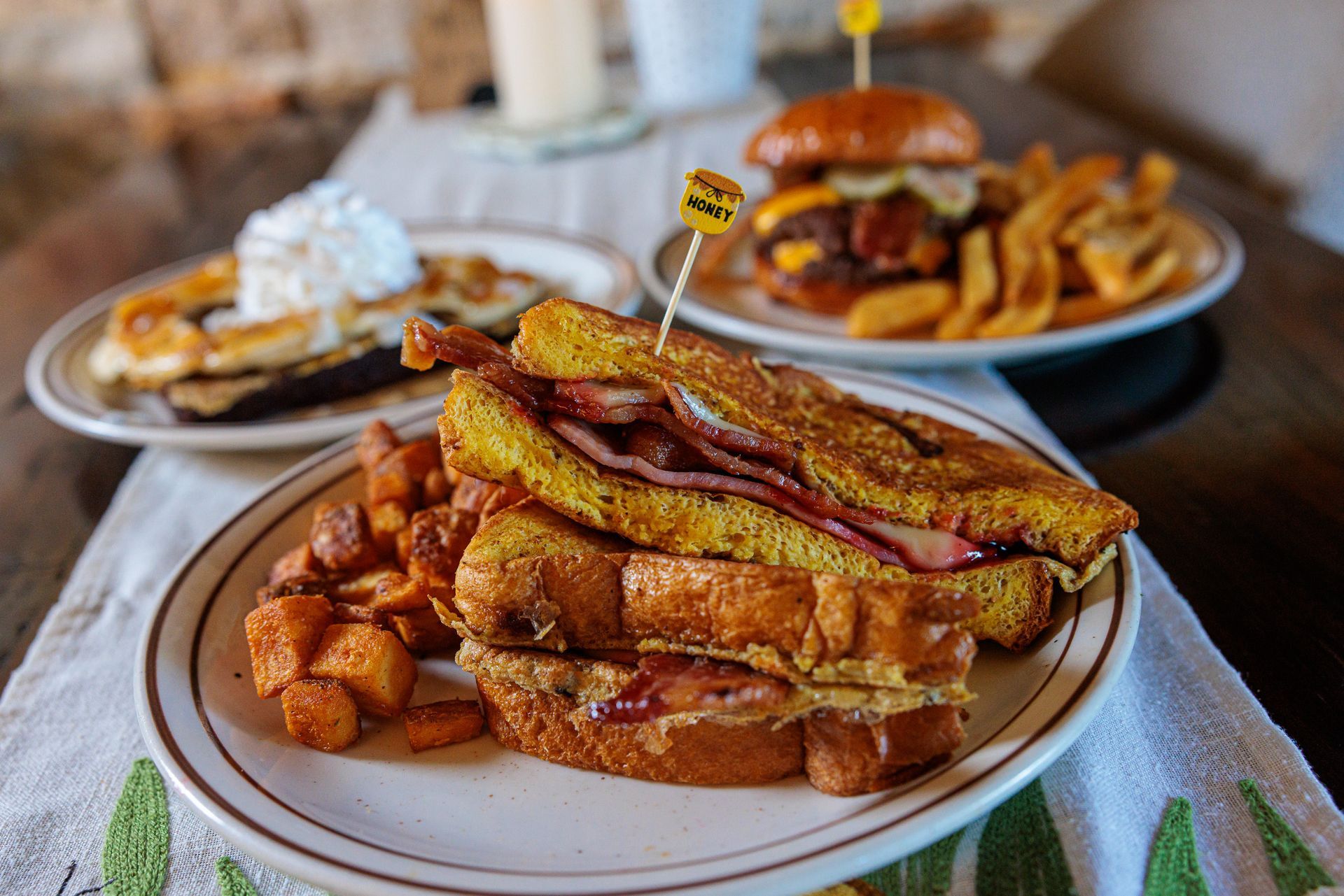 Plate of French toast sandwich, bacon, sweet potato cubes, with burger and dessert in background.