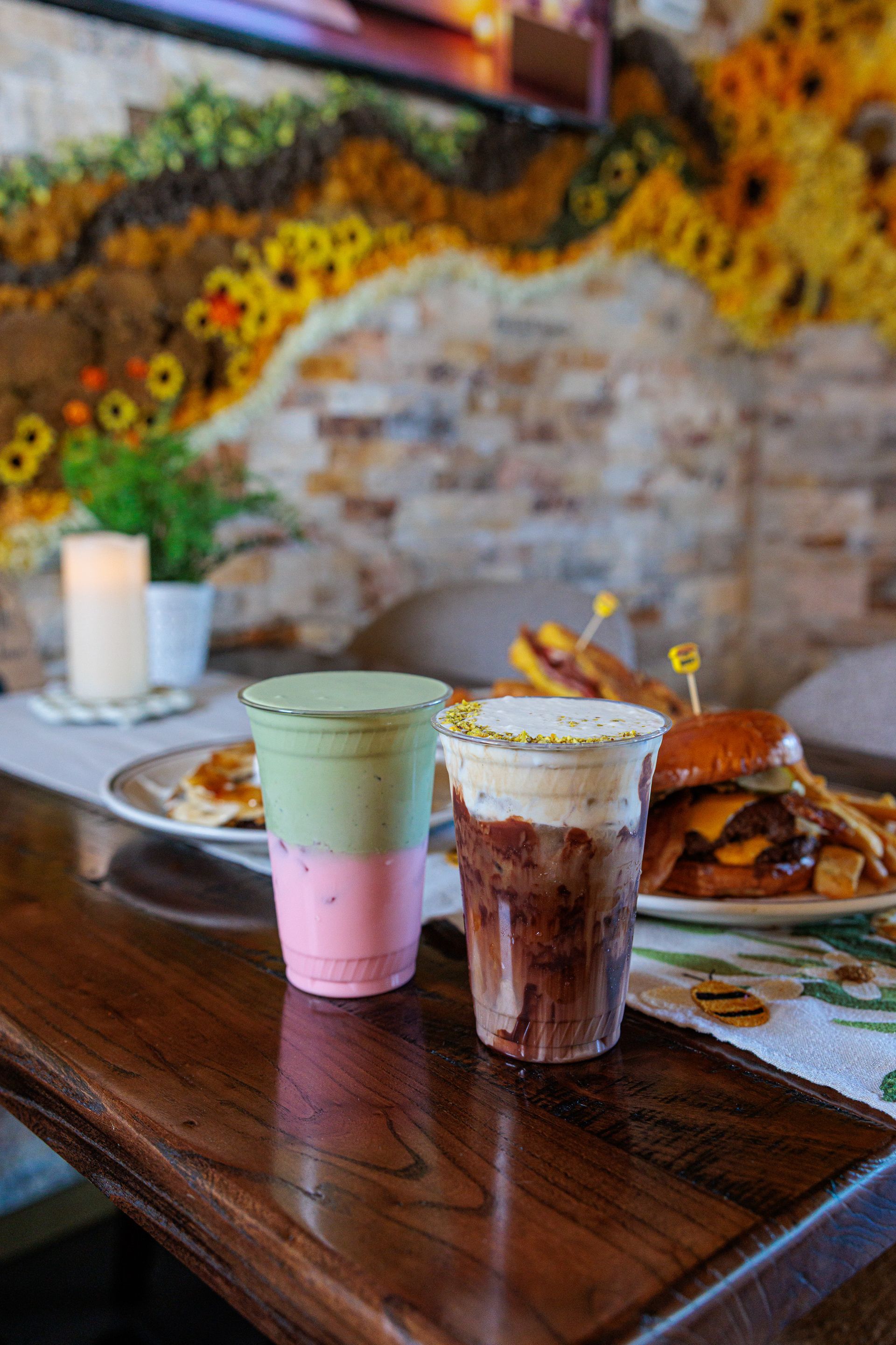 Two layered drinks on a wooden table, with food in the background. One is pink, green; the other is brown with whipped cream.