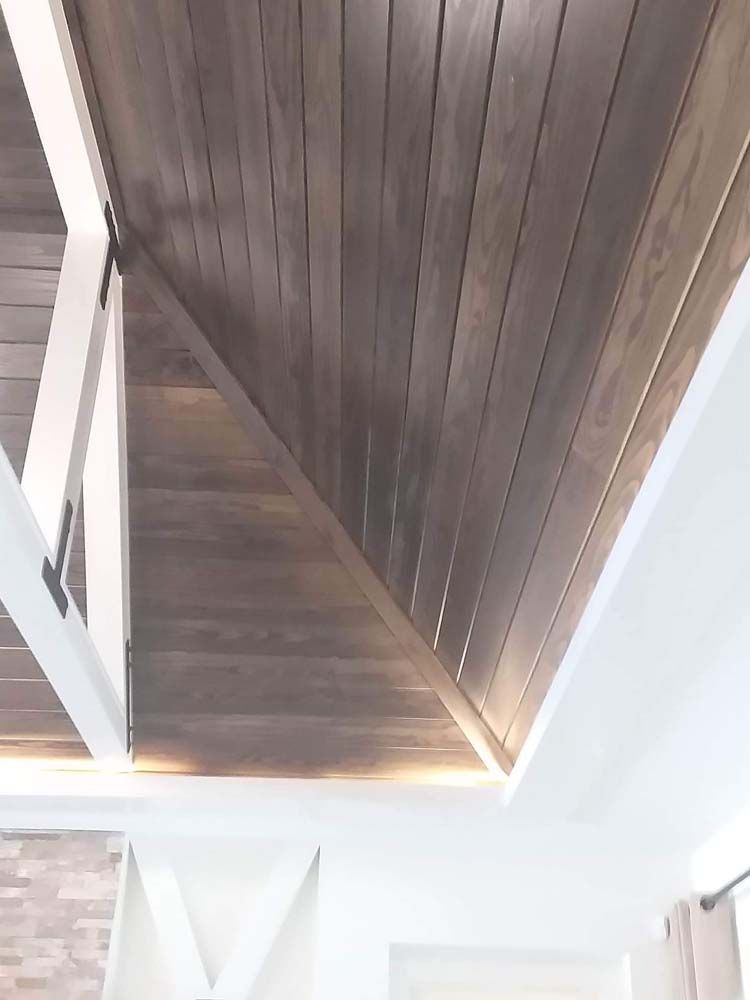 Wooden planks on an angled ceiling with recessed lighting and white beams.