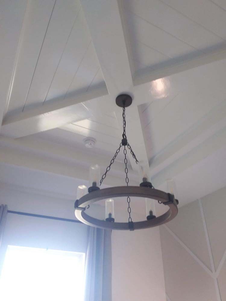 Chandelier hangs from a white wooden beamed ceiling. Circular wooden frame holds glass shades and lights.