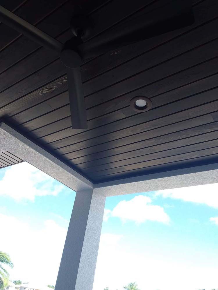 View from below of a dark wood-paneled ceiling with a fan and recessed light against a blue sky.