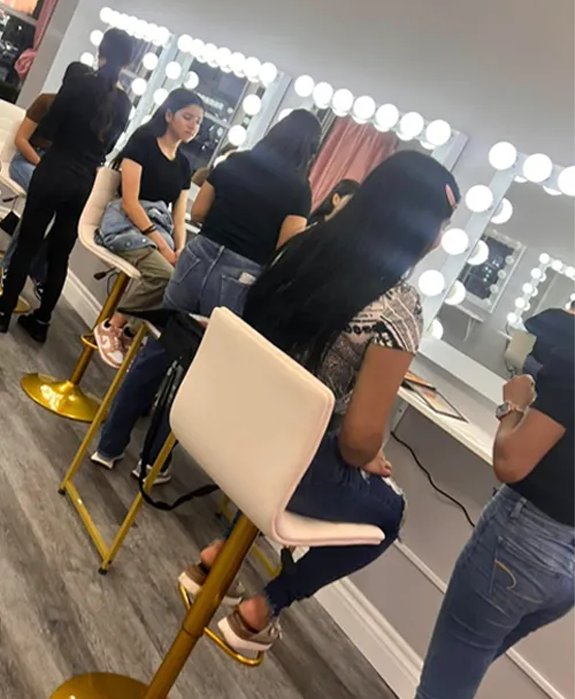 Makeup class with students sitting at stations in front of illuminated mirrors. One student has long black hair.
