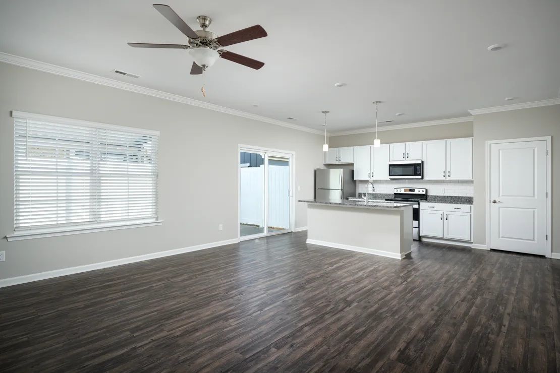 An empty living room with a ceiling fan and a kitchen in the background.
