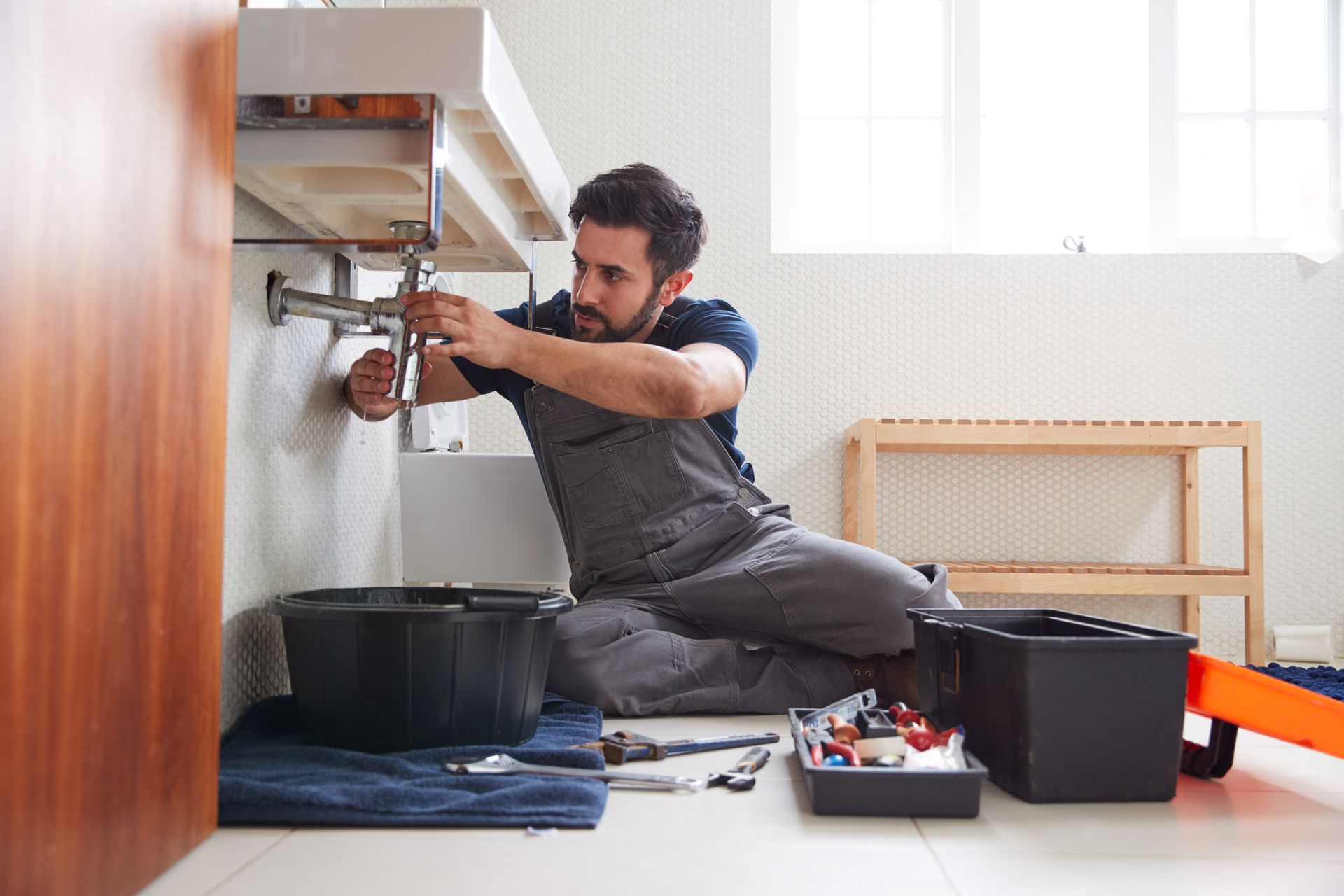 Plumber kneeling, fixing pipes under a white bathroom sink, tools nearby.
