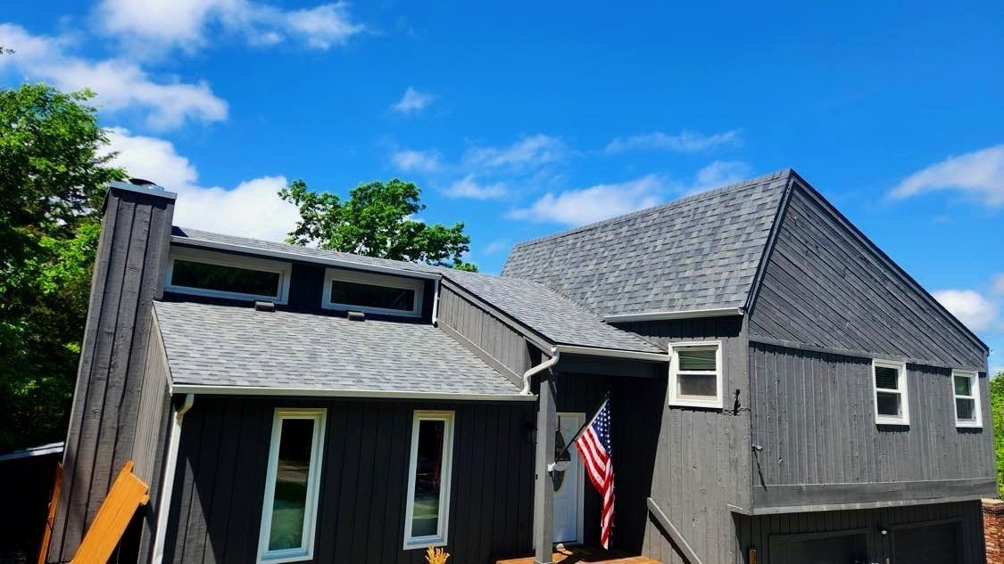 Gray house with dark roof, white-trim windows, and an American flag under a bright blue sky.