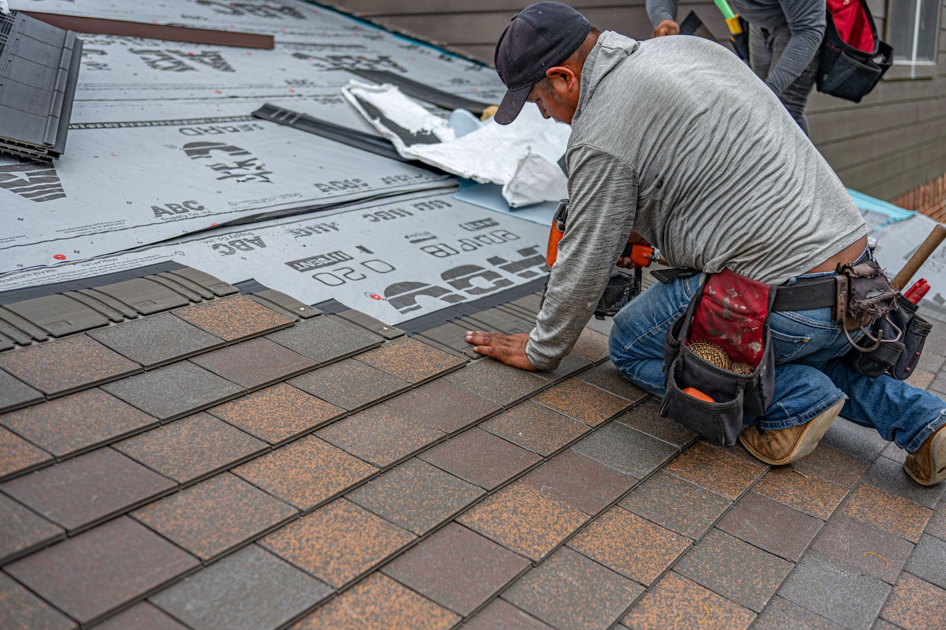Worker installing brown roof shingles beside gray underlayment on a rooftop