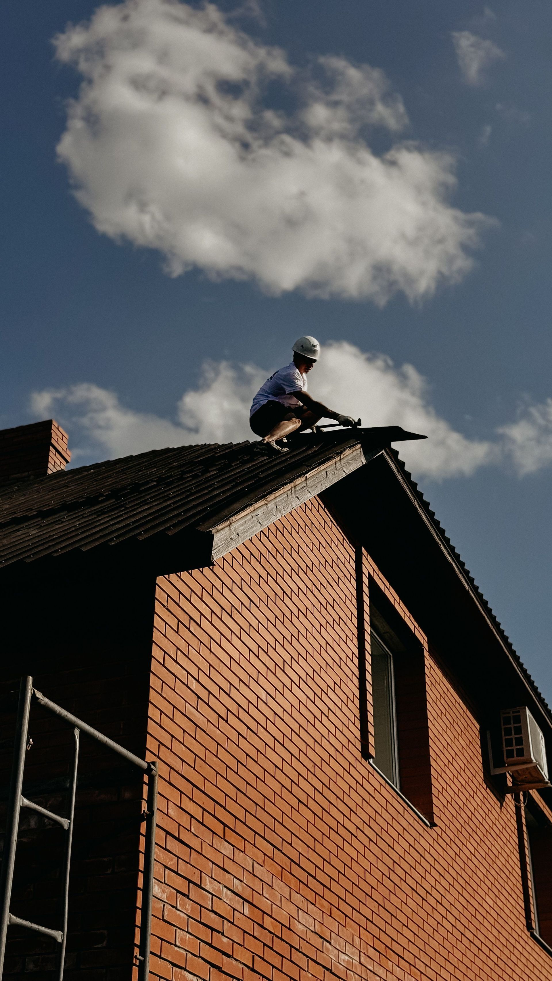 Worker on a brick roof under a cloudy sky, near a rooftop edge and chimney scaffolding.