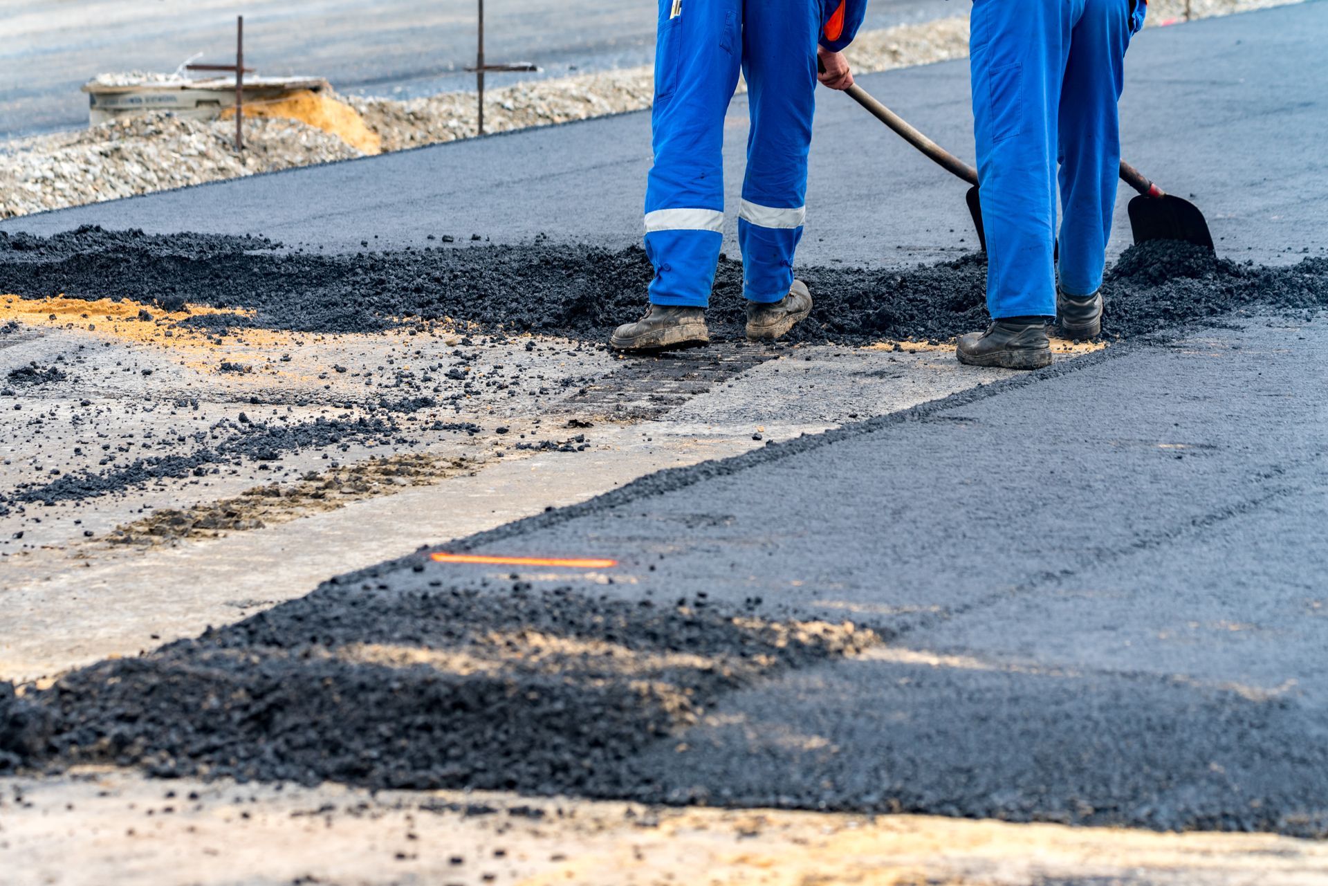 Two workers in blue coveralls paving a road with shovels; daytime.