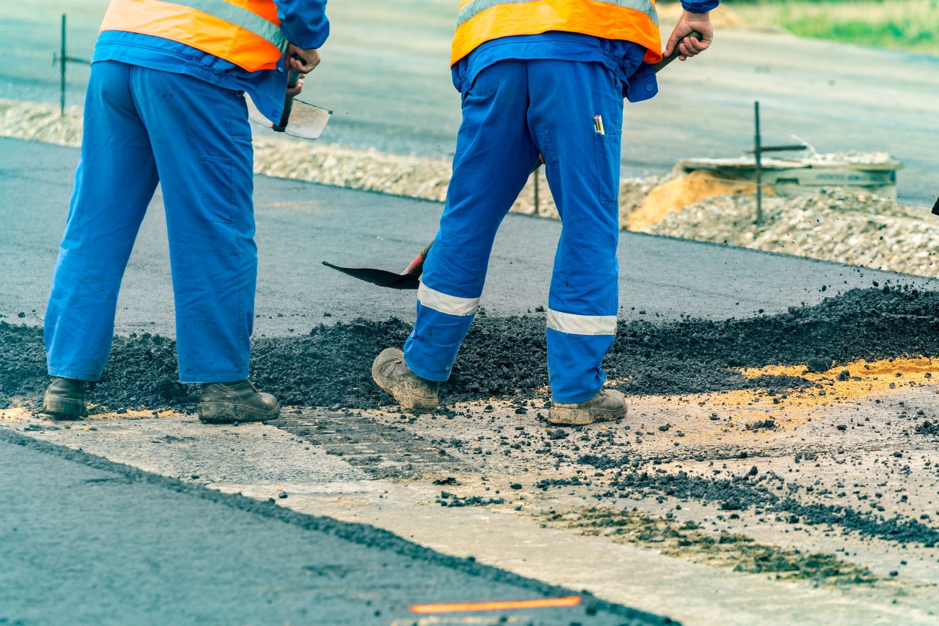 Two road workers in blue overalls and orange vests, laying asphalt on a street.