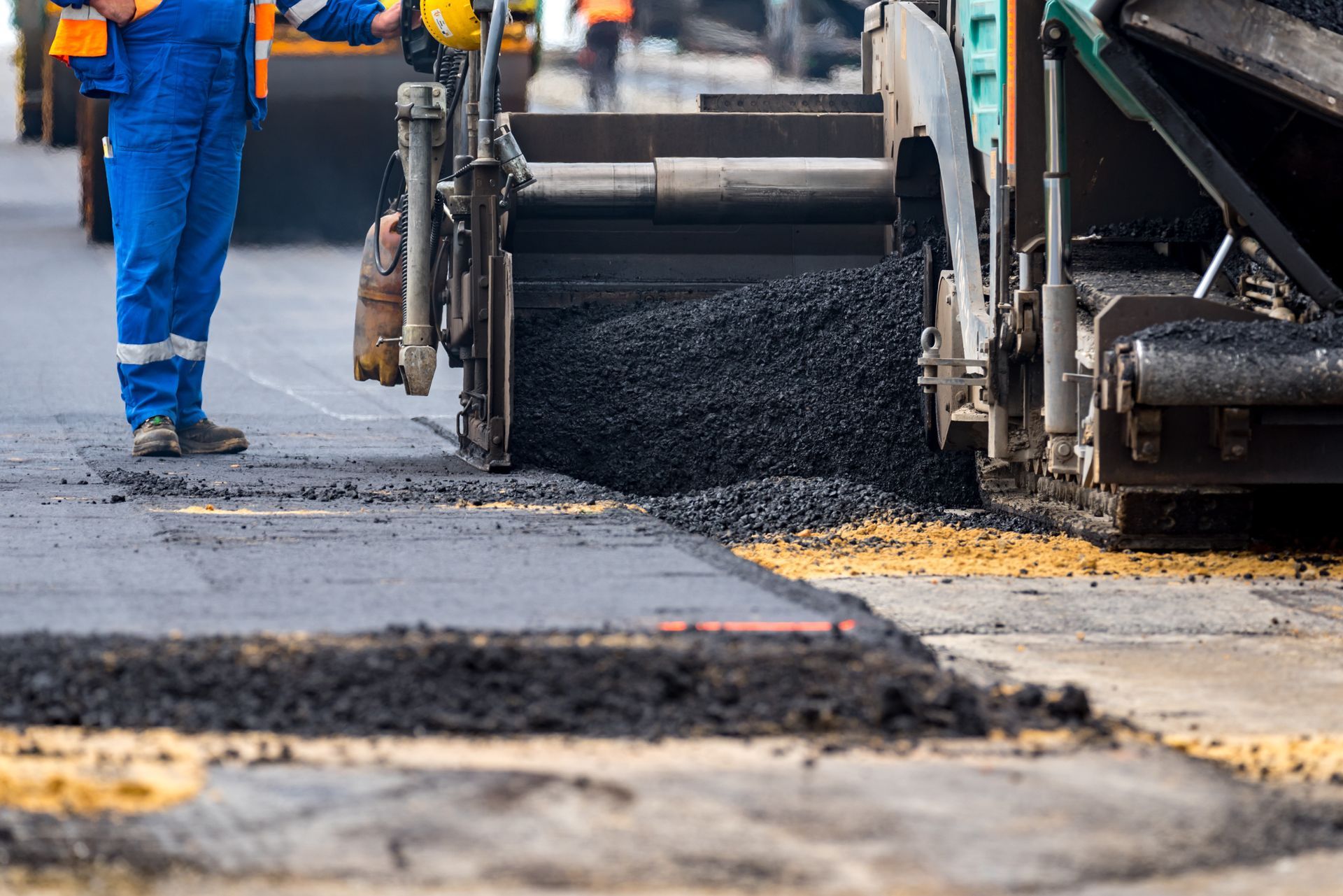 Road paving machine dispensing asphalt onto a road, construction worker in blue overalls nearby.