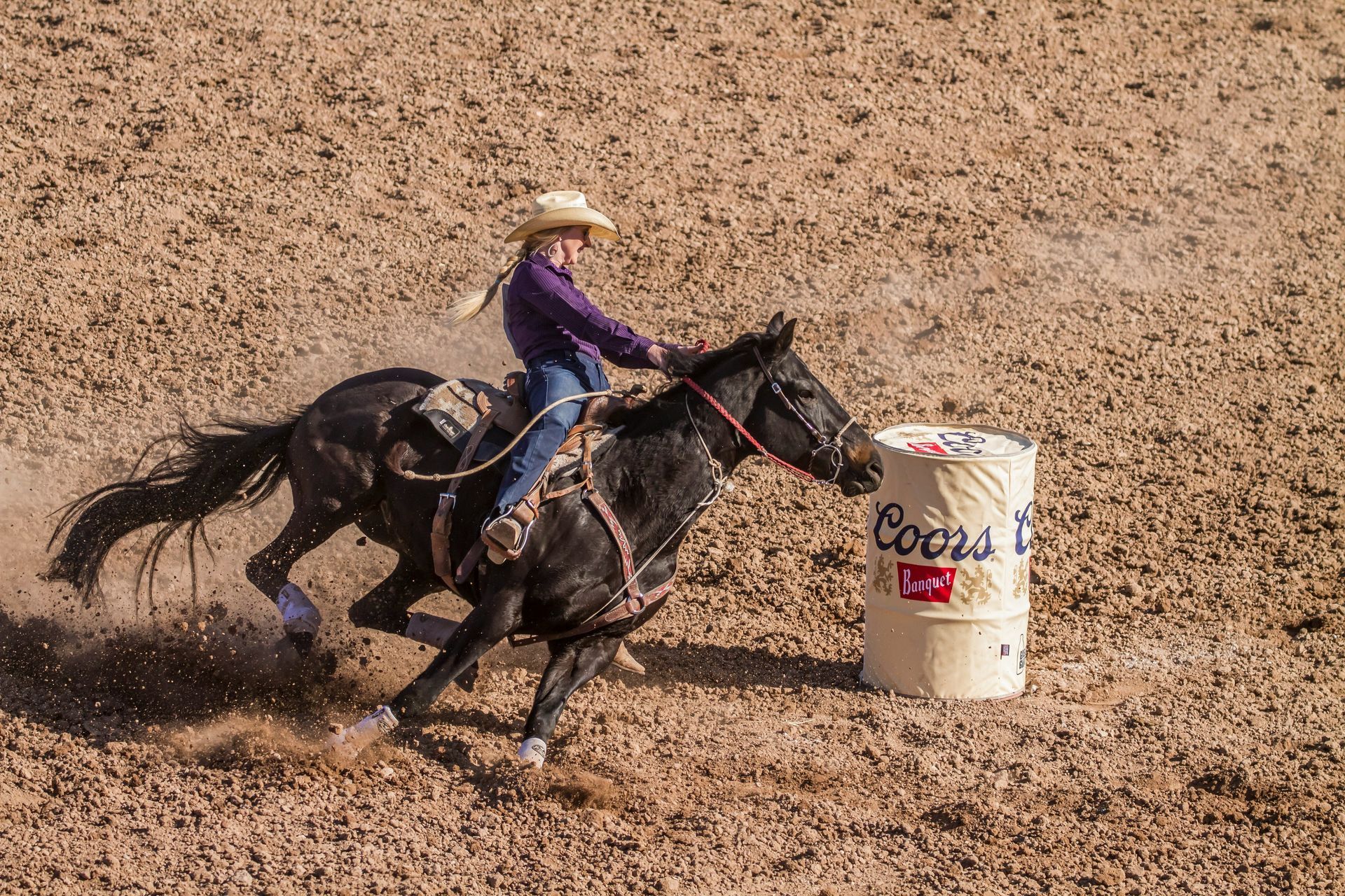 A young cowgirl in a rodeo, riding a black horse around a barrel, kicking up dirt.
