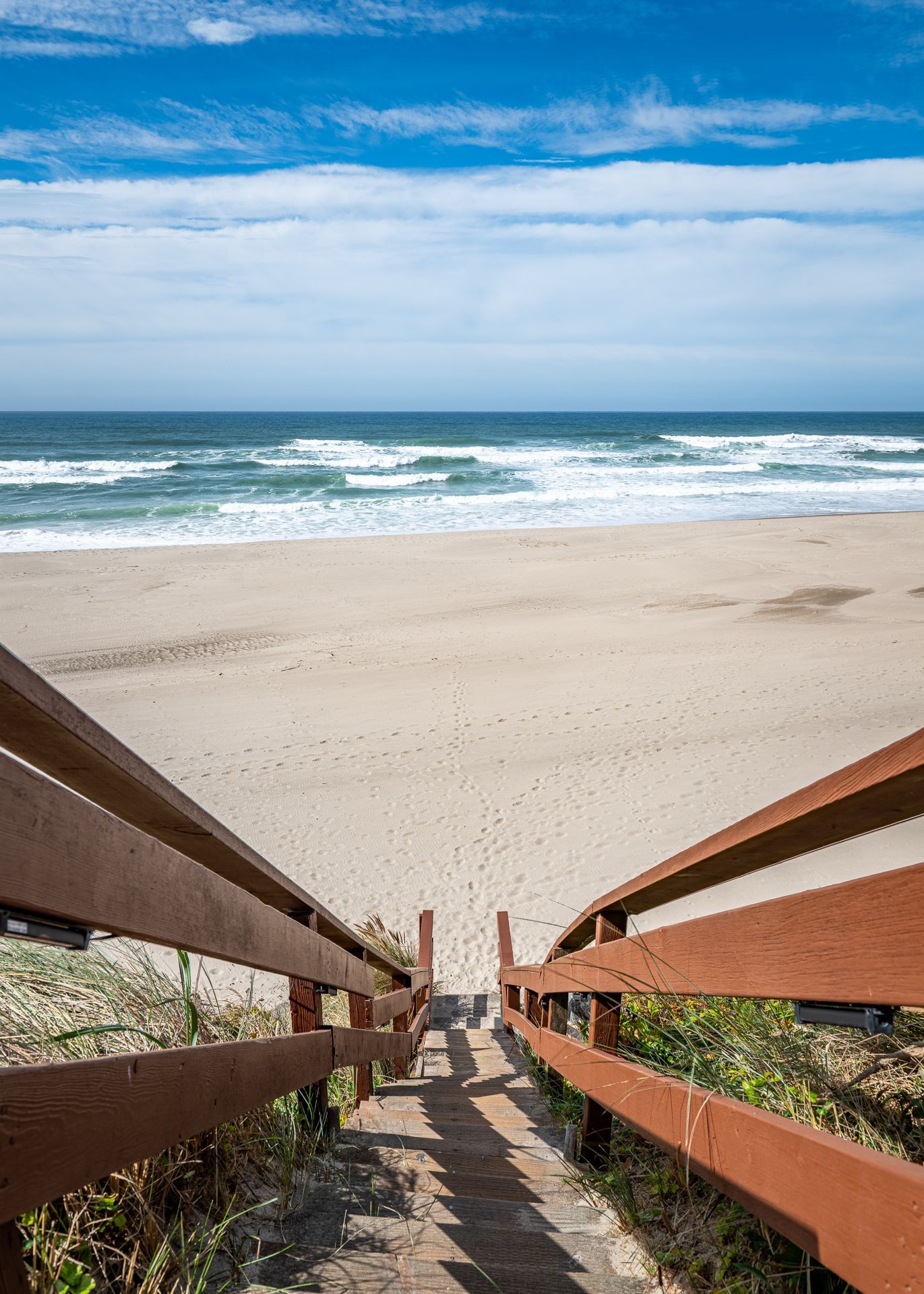 A wooden staircase leads down through grassy dunes to a sandy beach and the blue ocean under a bright, cloudy sky.