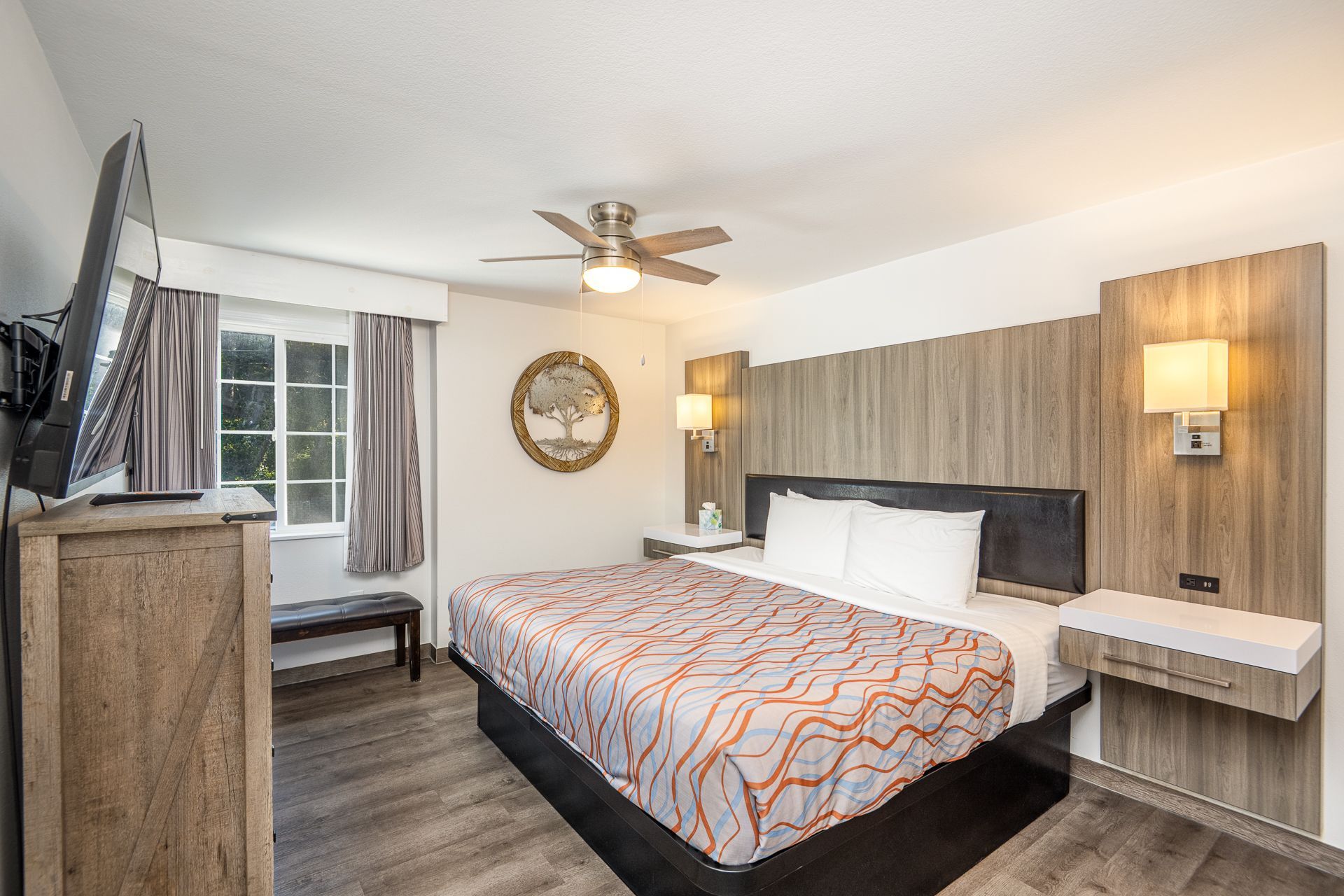 A hotel room with a queen bed, wood-paneled headboard, floating nightstands, and a large television on a wood dresser.