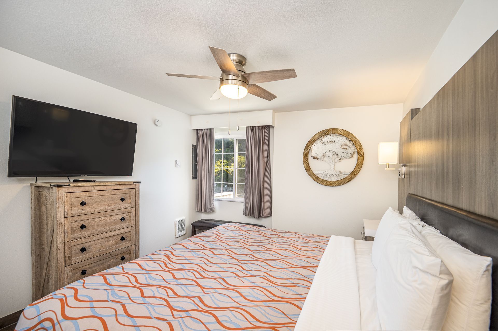 A bedroom with a bed featuring an orange-patterned quilt, a rustic wooden dresser with a TV, and a circular wall decor.