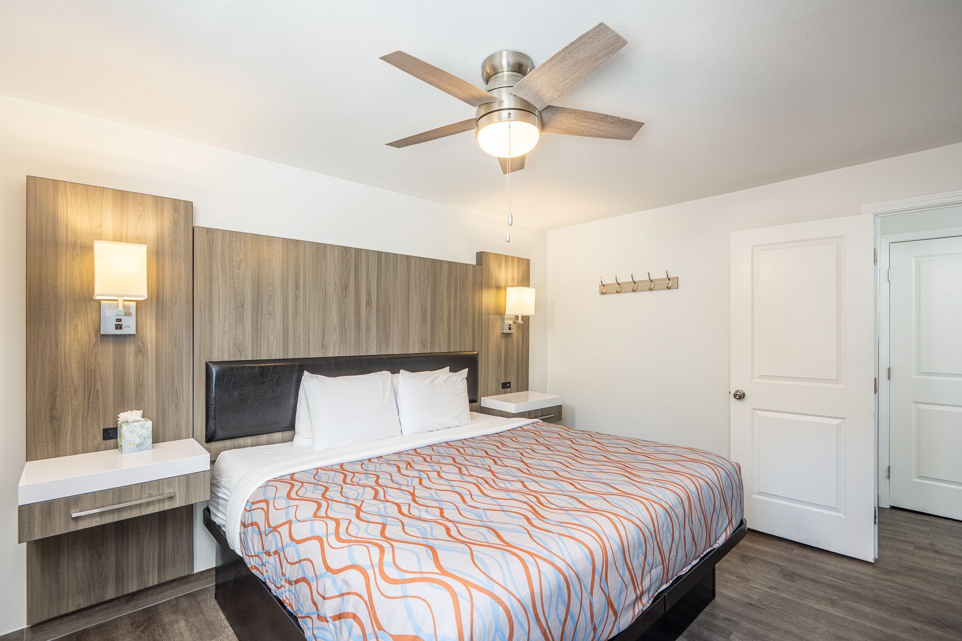 A modern hotel room with a large bed, light wood headboard, floating nightstands, and a ceiling fan.
