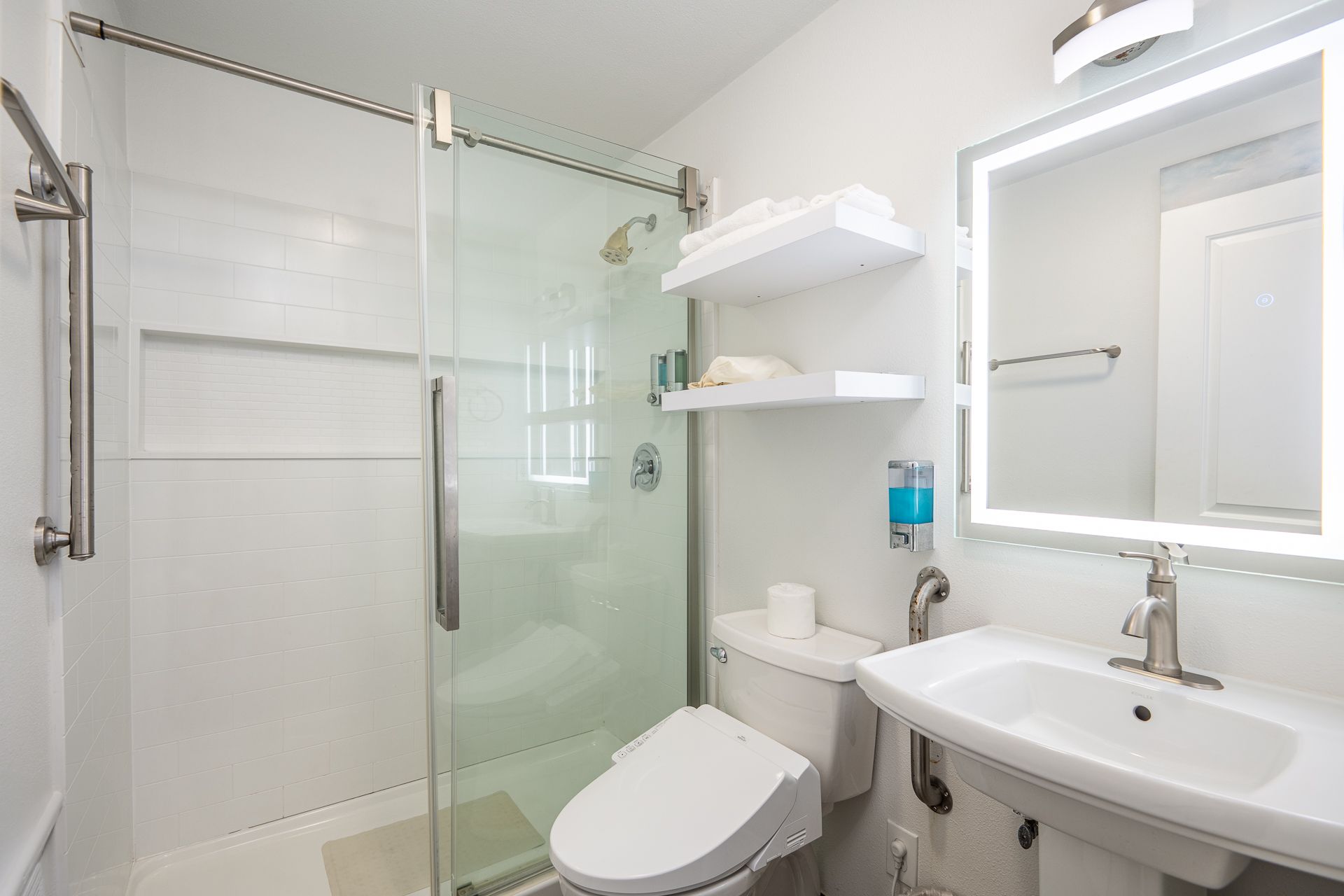 A bright, accessible bathroom with a glass-enclosed shower, wall shelves, a toilet, and a sink with an illuminated mirror.