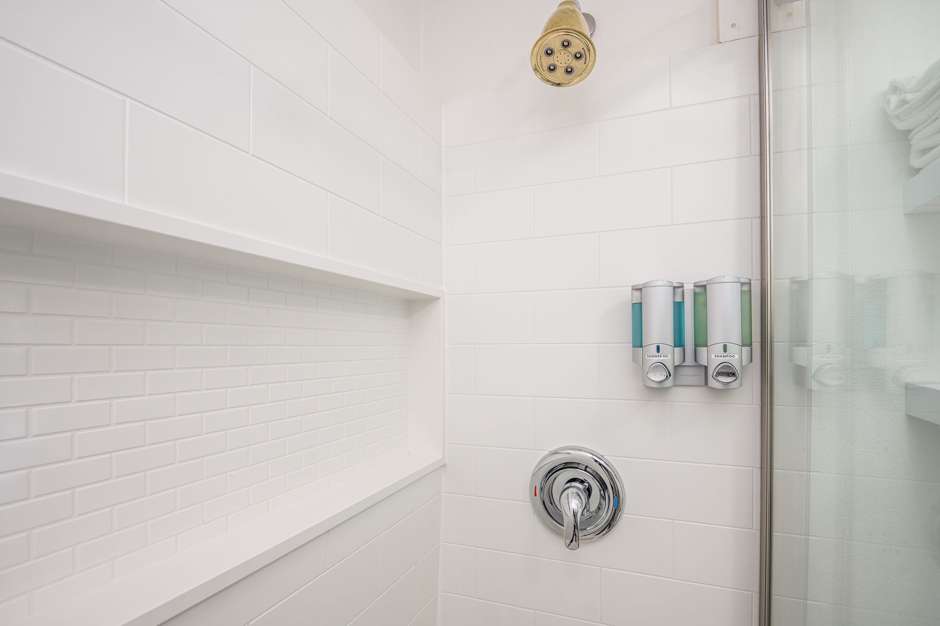 A white tiled shower with a chrome shower head, a control handle, and wall-mounted soap dispensers.