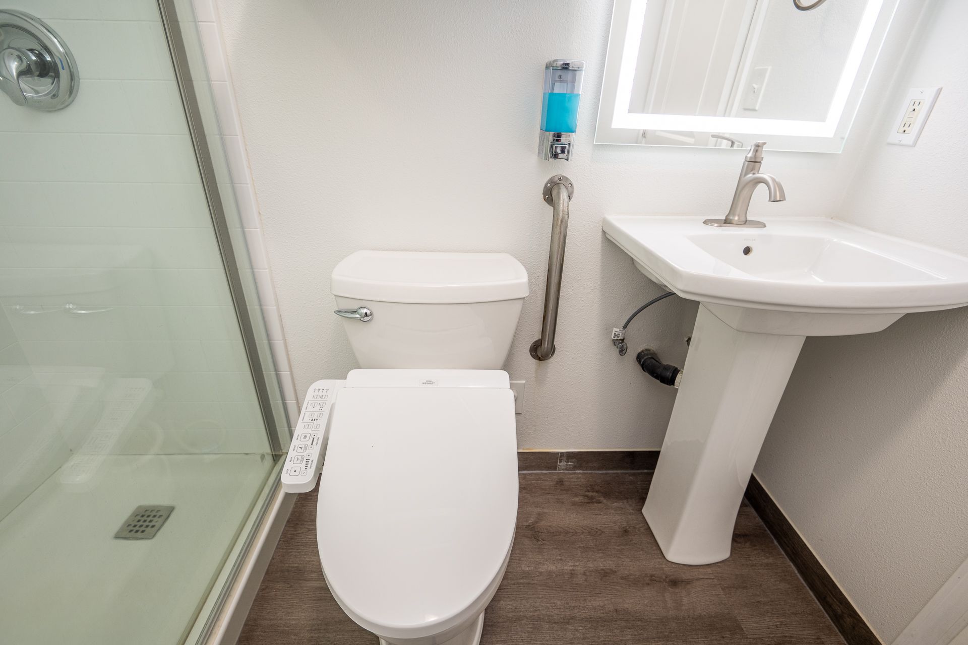 An accessible bathroom with a white pedestal sink, a toilet with a bidet seat, and a vertical grab bar on the wall.