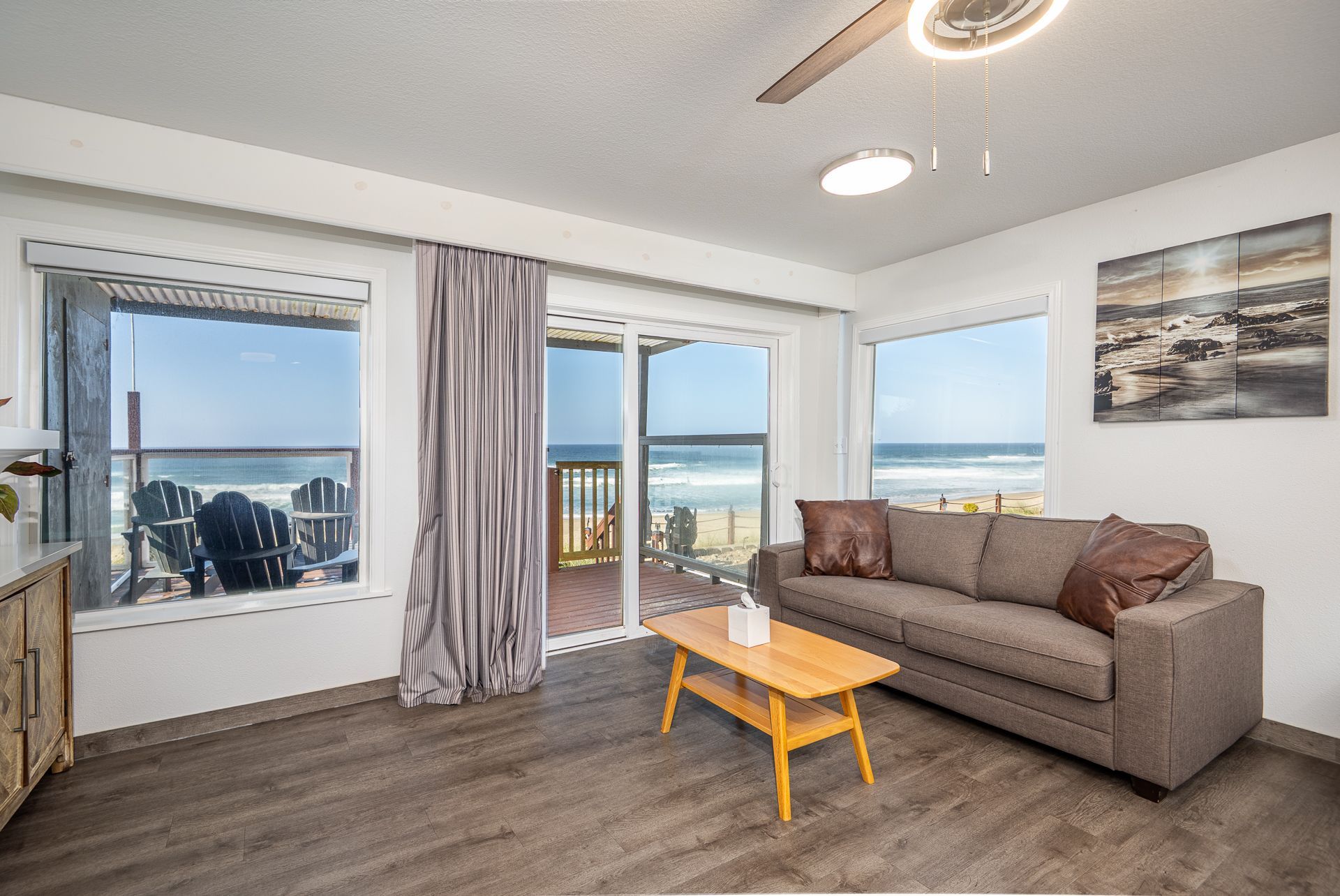 A modern living room with a gray sofa, wood coffee table, and large windows overlooking a sunny beach and ocean.