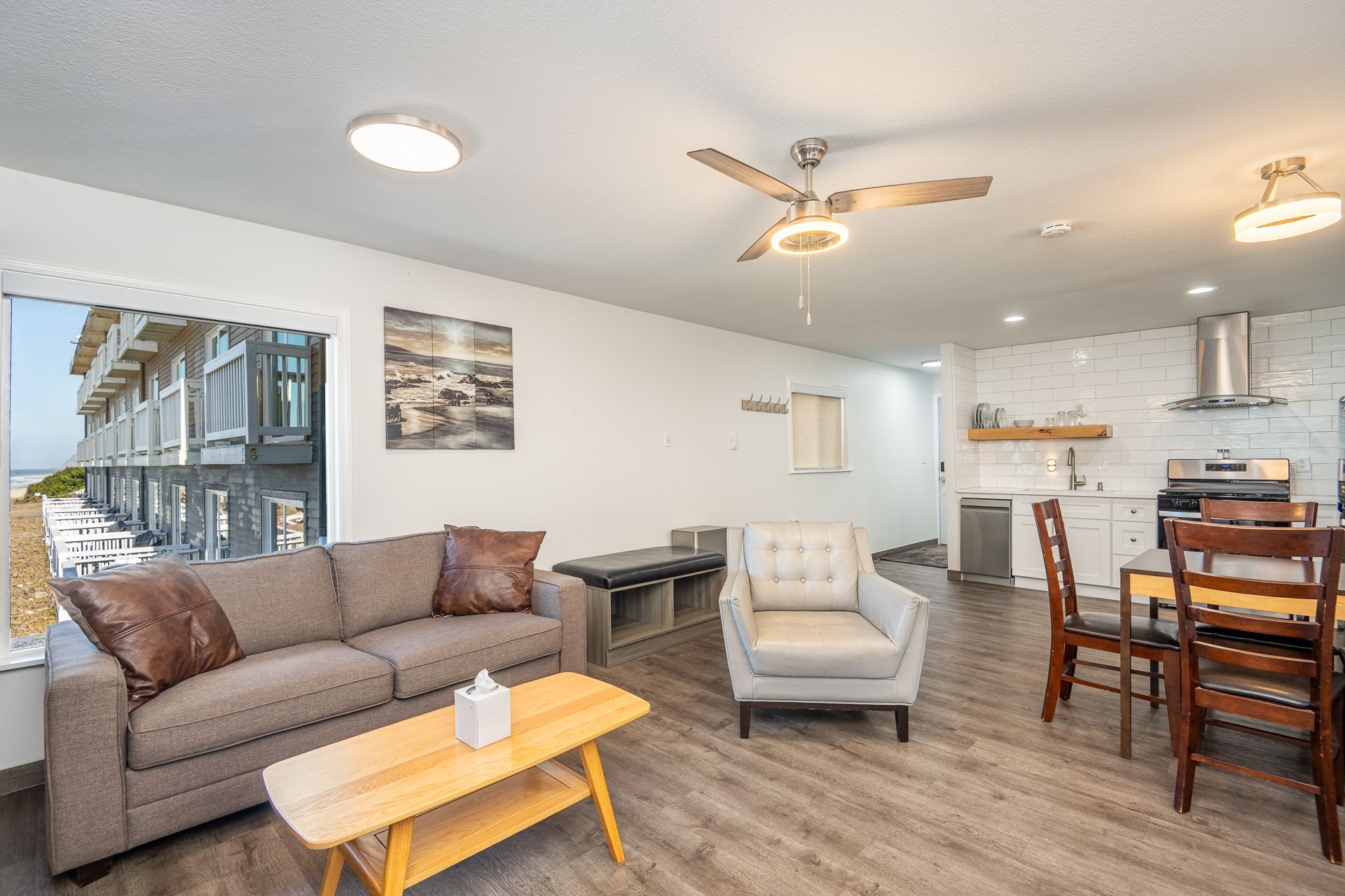 A modern living room with a brown couch, white armchair, wooden coffee table, and an open kitchen with dining table.
