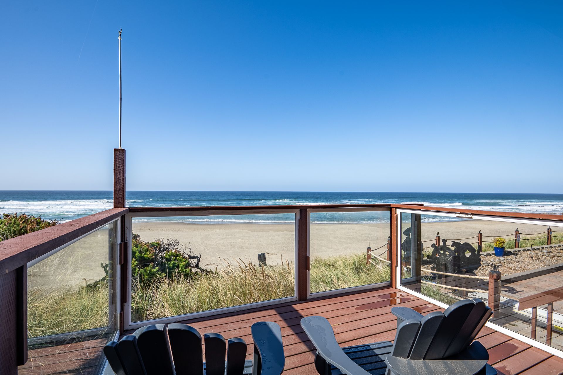 Two Adirondack chairs sit on a wooden deck overlooking a sandy beach and the ocean under a clear blue sky.