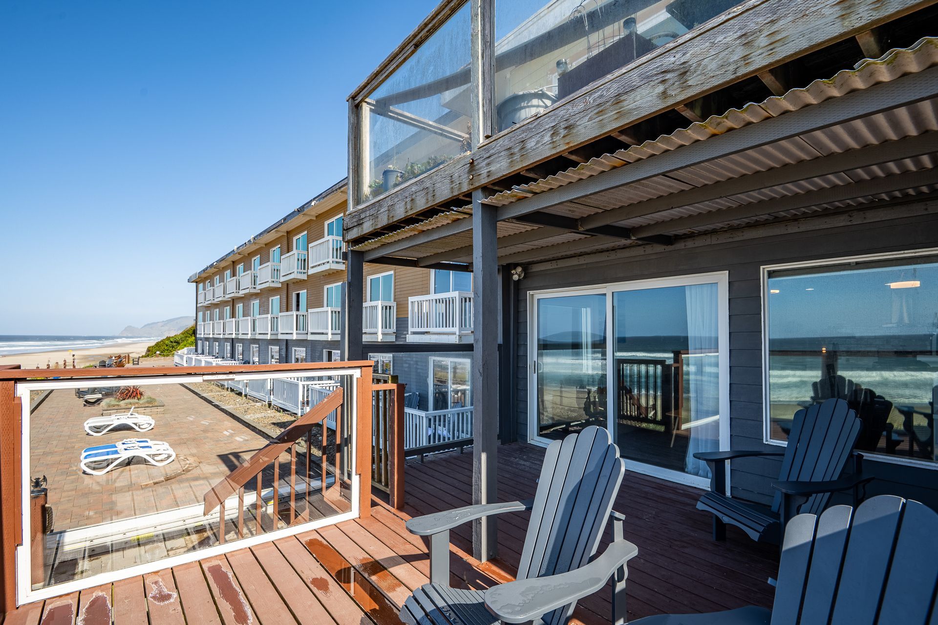 A balcony with grey wooden chairs overlooks a beach hotel and the ocean on a sunny day.