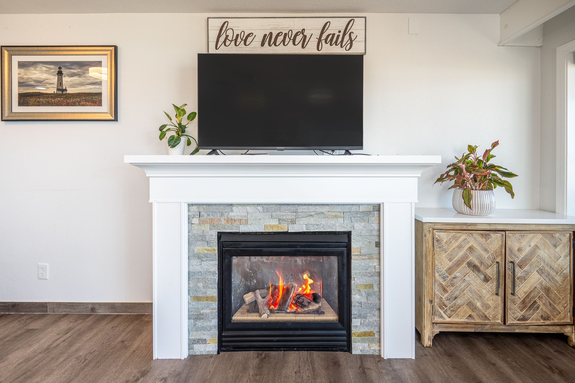 A fireplace with stone tiling and a white mantle, featuring a television, house plants, a framed picture, and a cabinet.