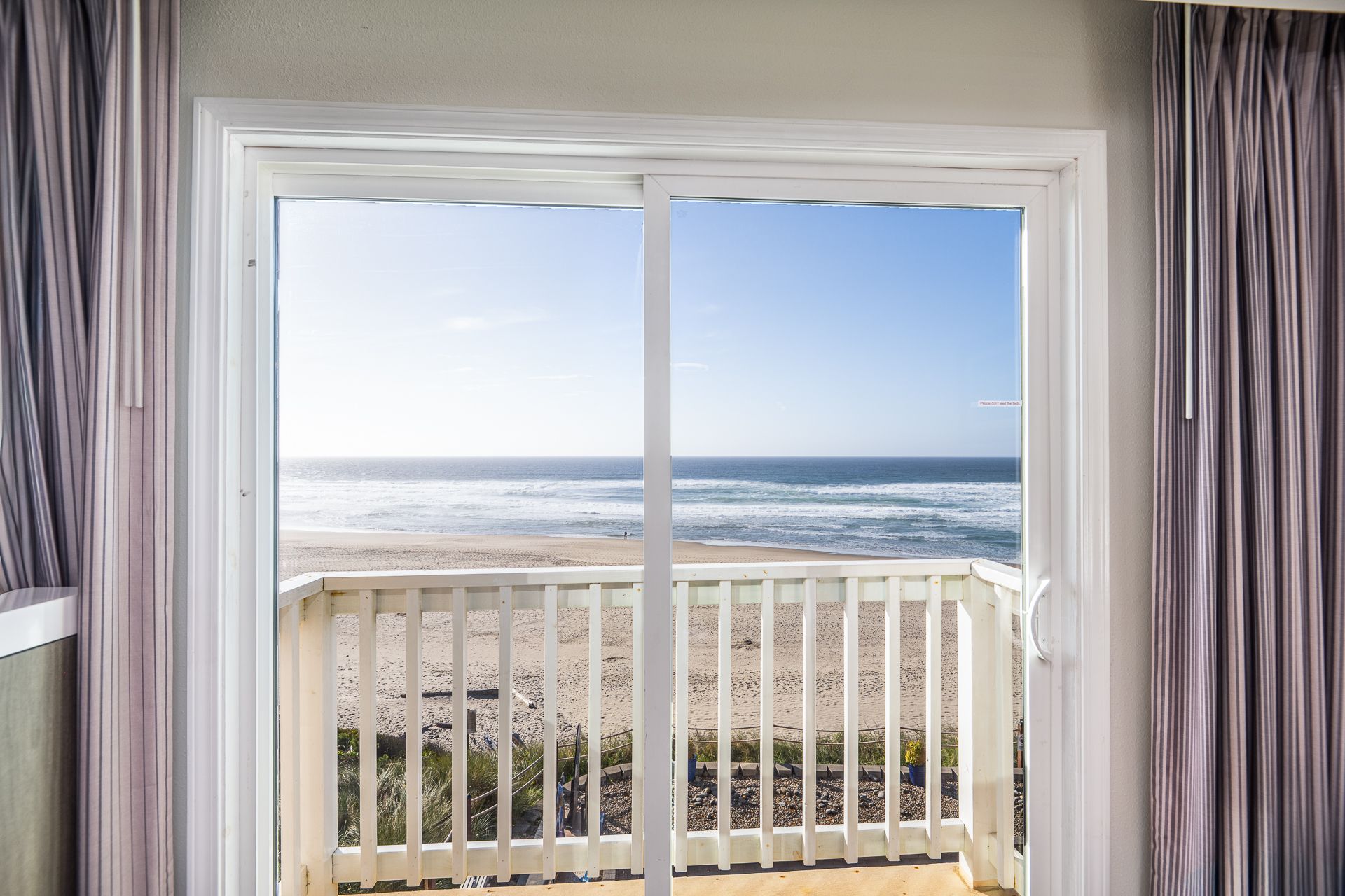 A view of a sandy beach and ocean through a sliding glass door, flanked by light purple vertical curtains.