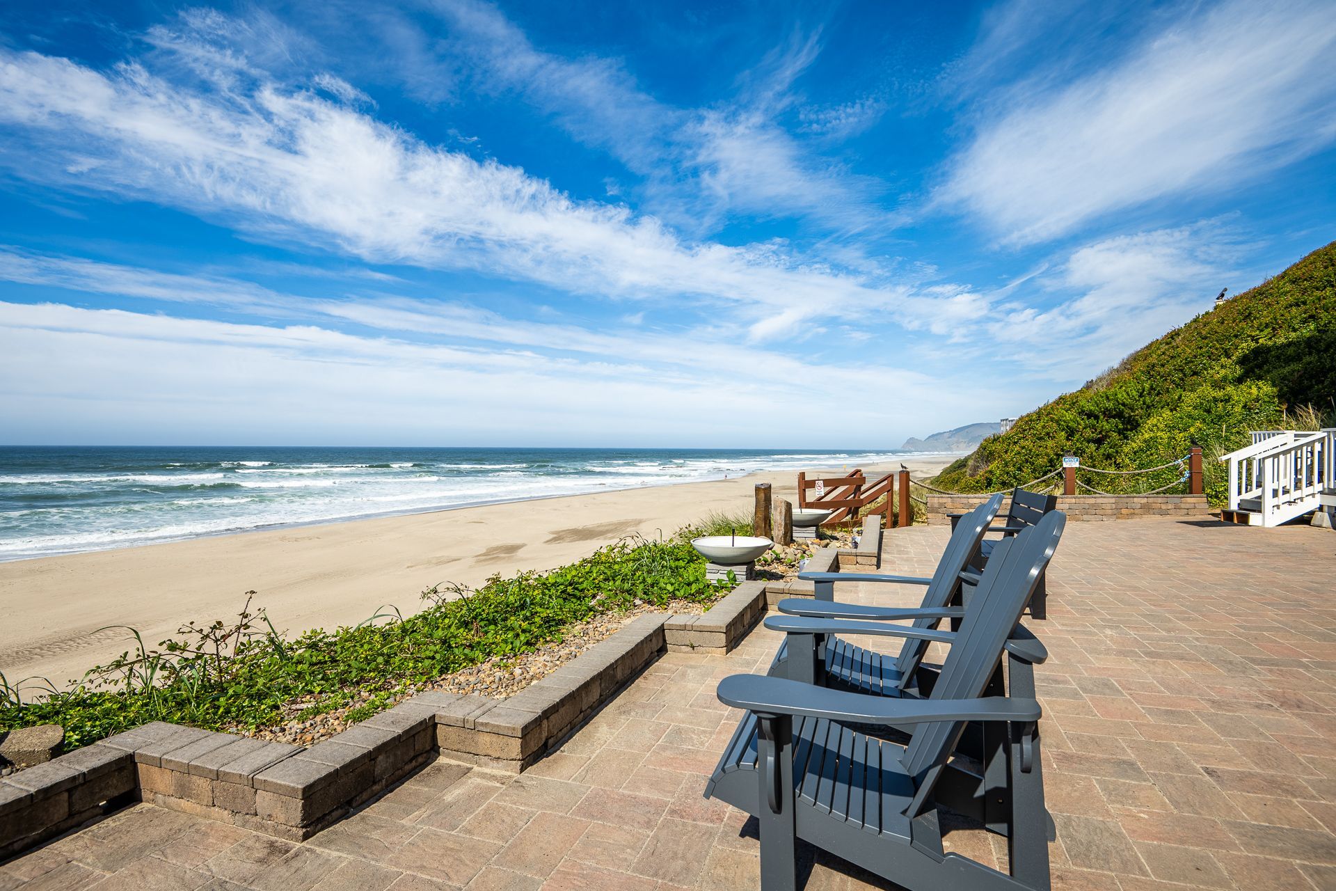 Two dark blue Adirondack chairs on a paved patio overlooking a sunny, sandy beach and the ocean under a blue sky.