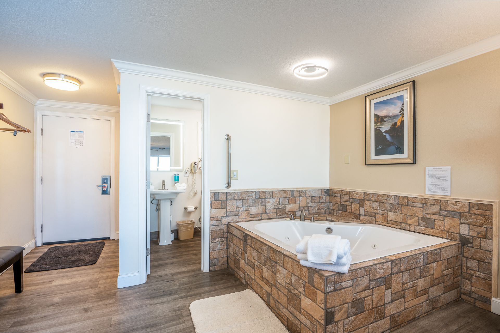 A hotel room featuring a corner stone-tiled jetted tub, an open bathroom doorway with a pedestal sink, and a white door.