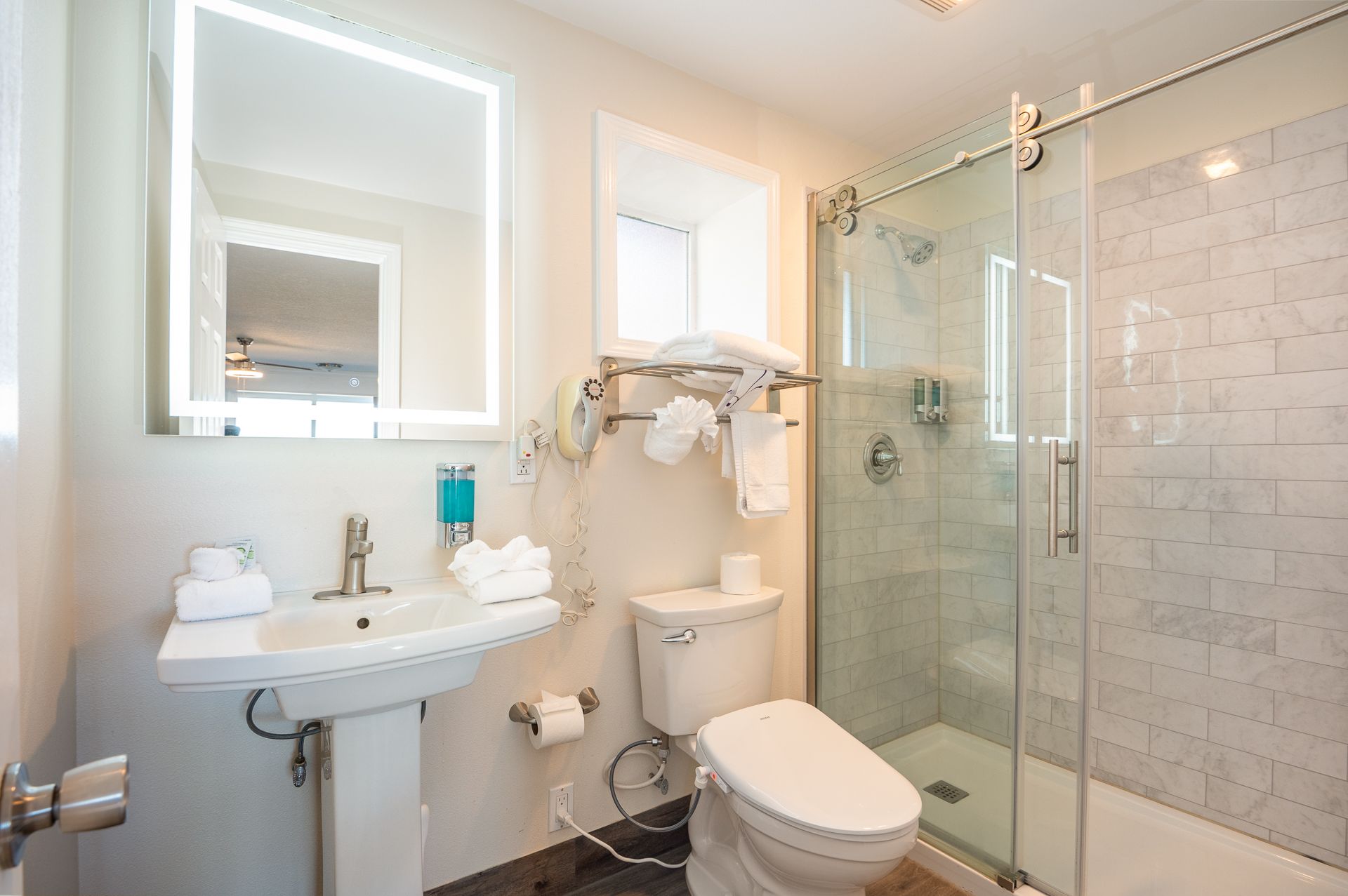 A bright, modern bathroom featuring a pedestal sink, white toilet, and a glass-enclosed shower with light tiled walls.