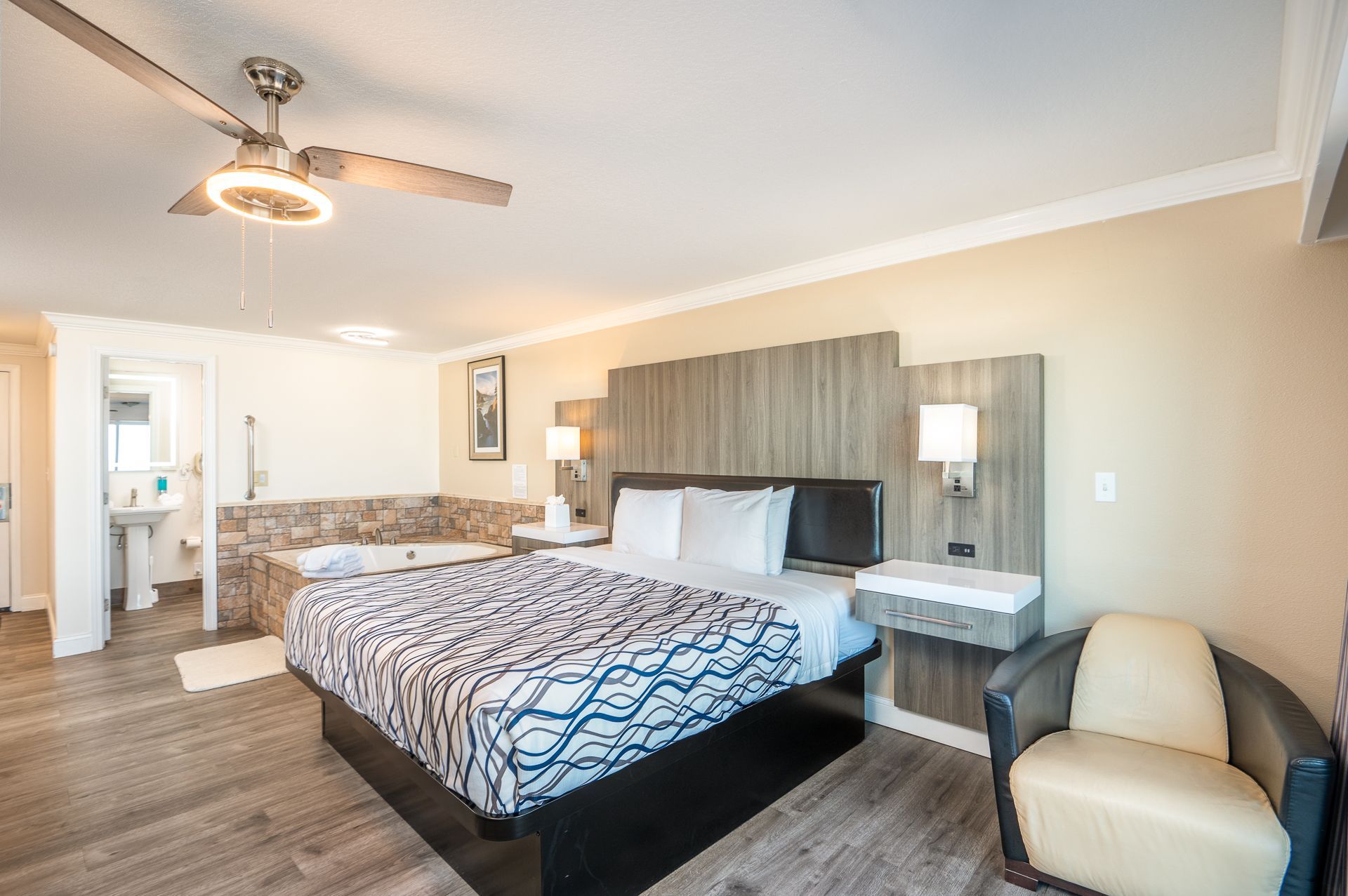 A hotel room with a queen bed featuring a blue and white patterned comforter, a ceiling fan, and a corner armchair.
