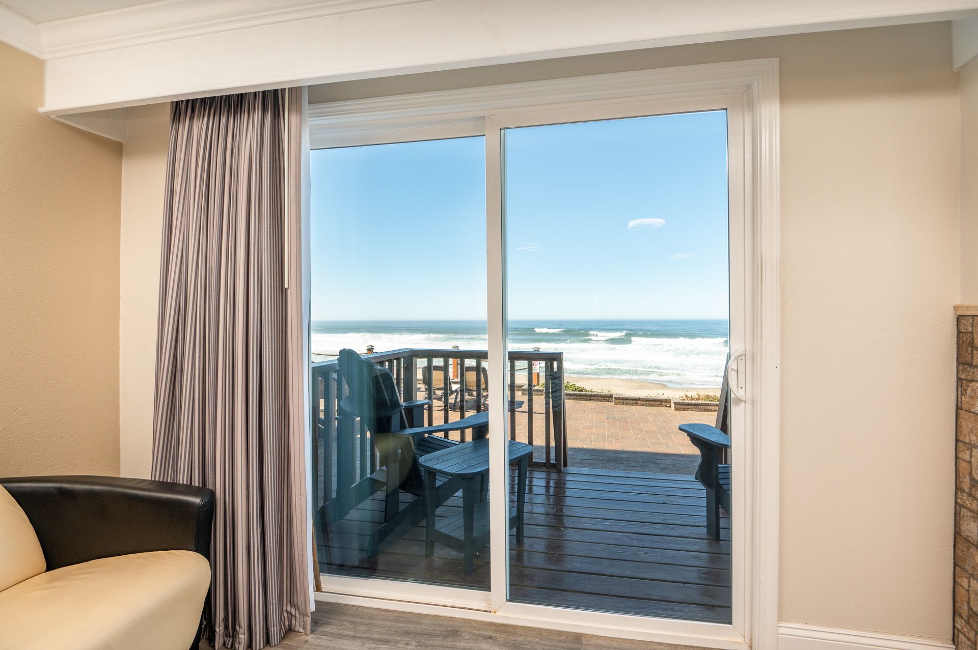 A view through a sliding glass door shows a balcony with two chairs overlooking a sandy beach and the ocean.