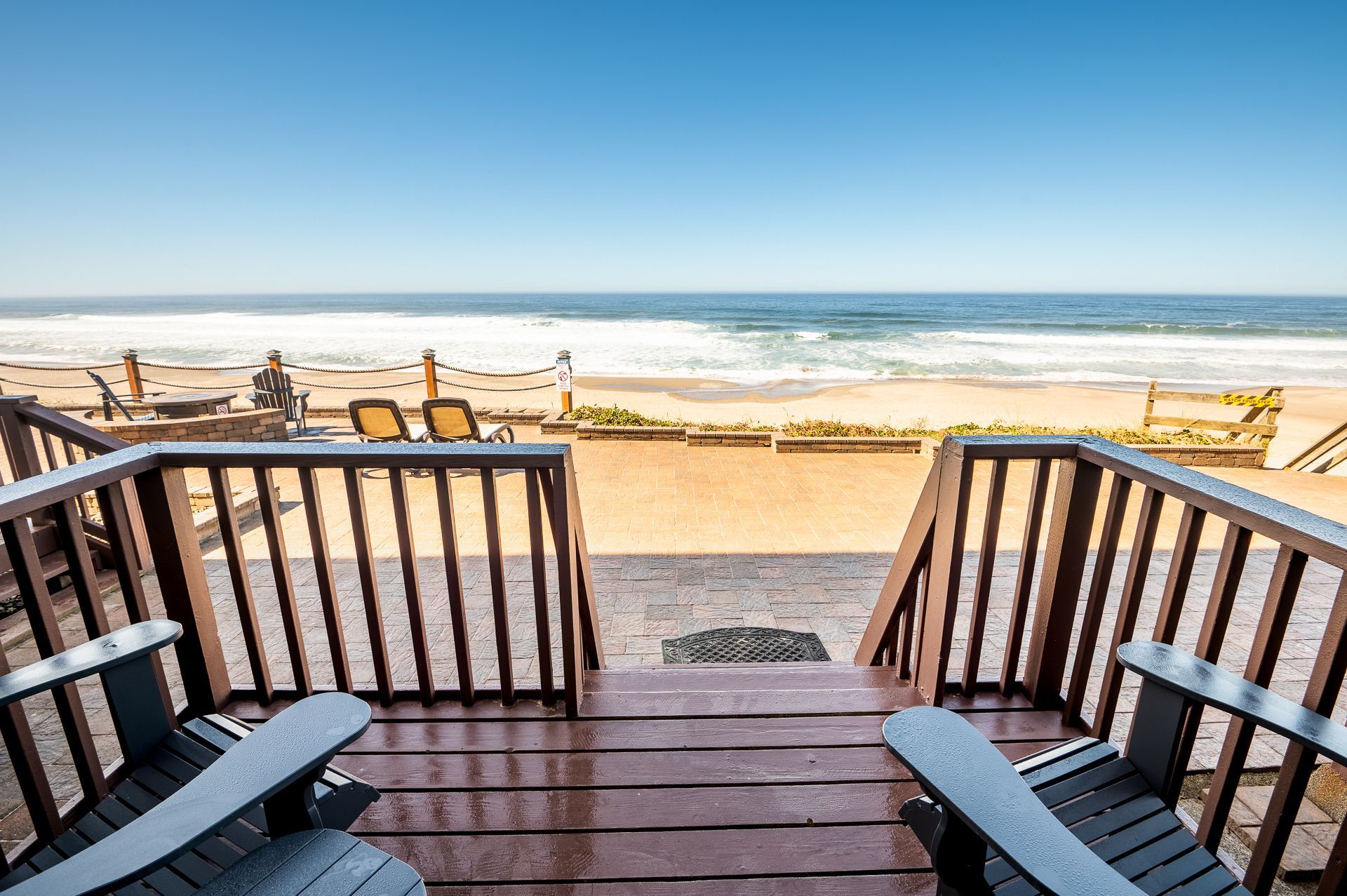 A view from a wooden deck with two chairs, overlooking a sandy beach and the ocean under a clear blue sky.