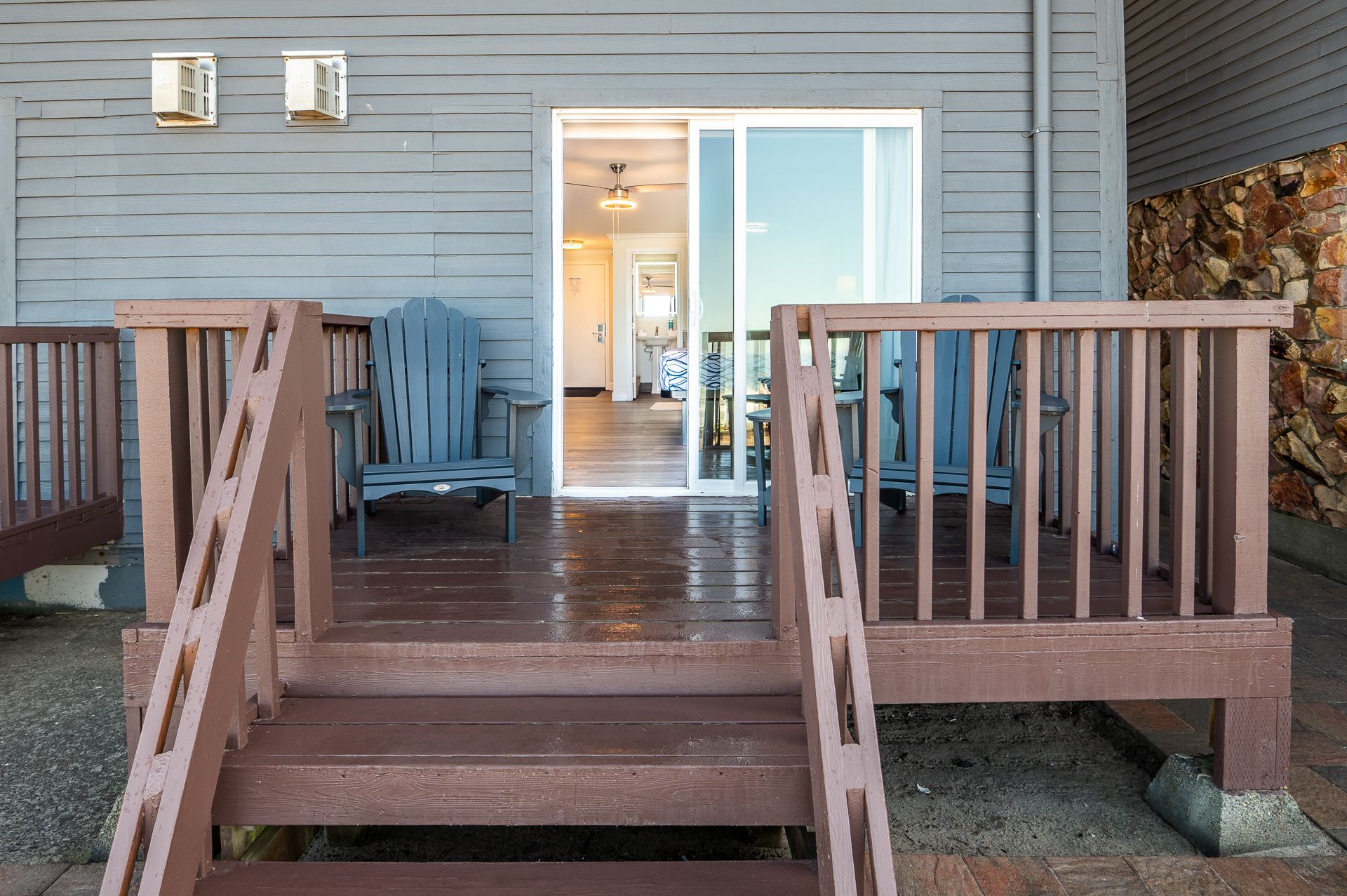 A gray-sided building exterior with a wooden deck, a sliding glass door, and two blue Adirondack chairs.