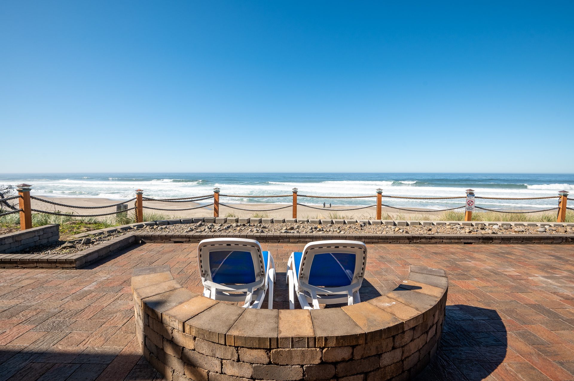 Two beach lounge chairs on a brick patio facing a wooden fence, a sandy beach, and the ocean under a clear blue sky.