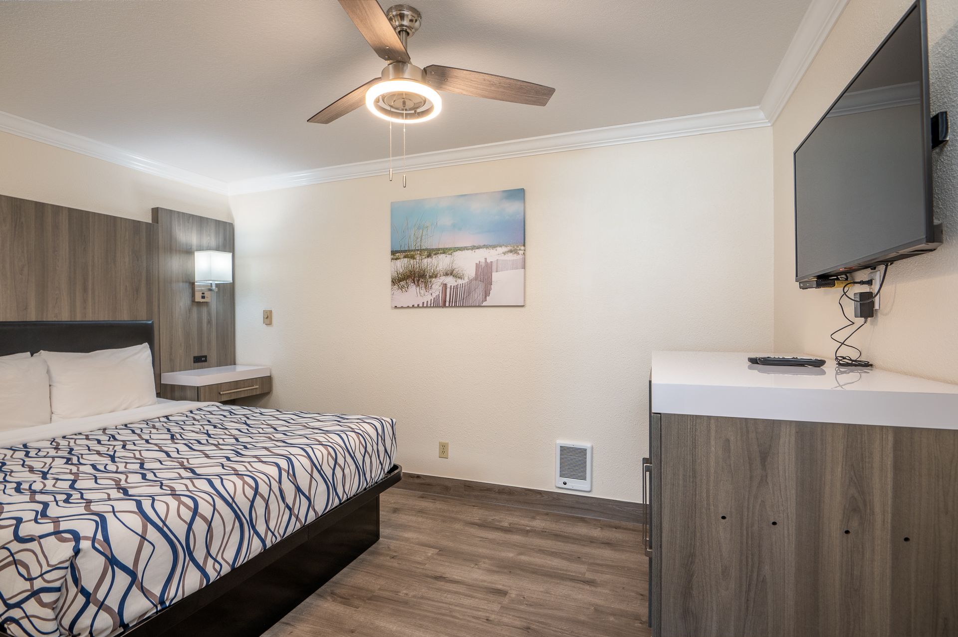 A hotel room with a queen bed featuring a blue patterned comforter, a wall-mounted TV, and a ceiling fan.