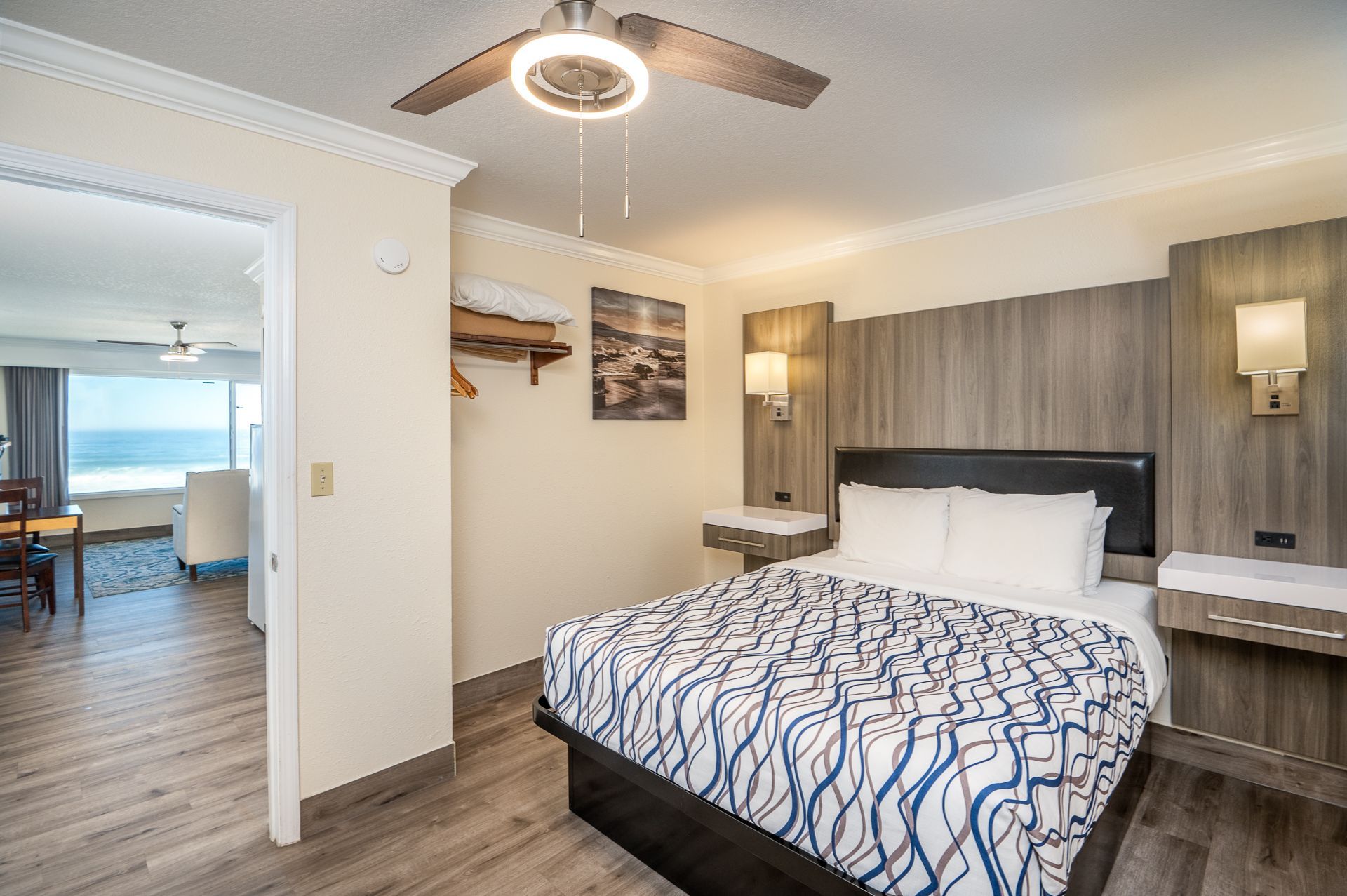 A hotel bedroom with a queen bed featuring blue patterned linens, wood paneling, and an adjacent view of the ocean.