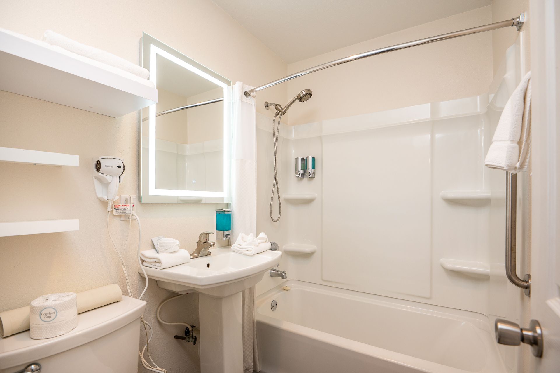 A bright, clean hotel bathroom with a white pedestal sink, toilet, tub-shower combination, and a lit mirror.