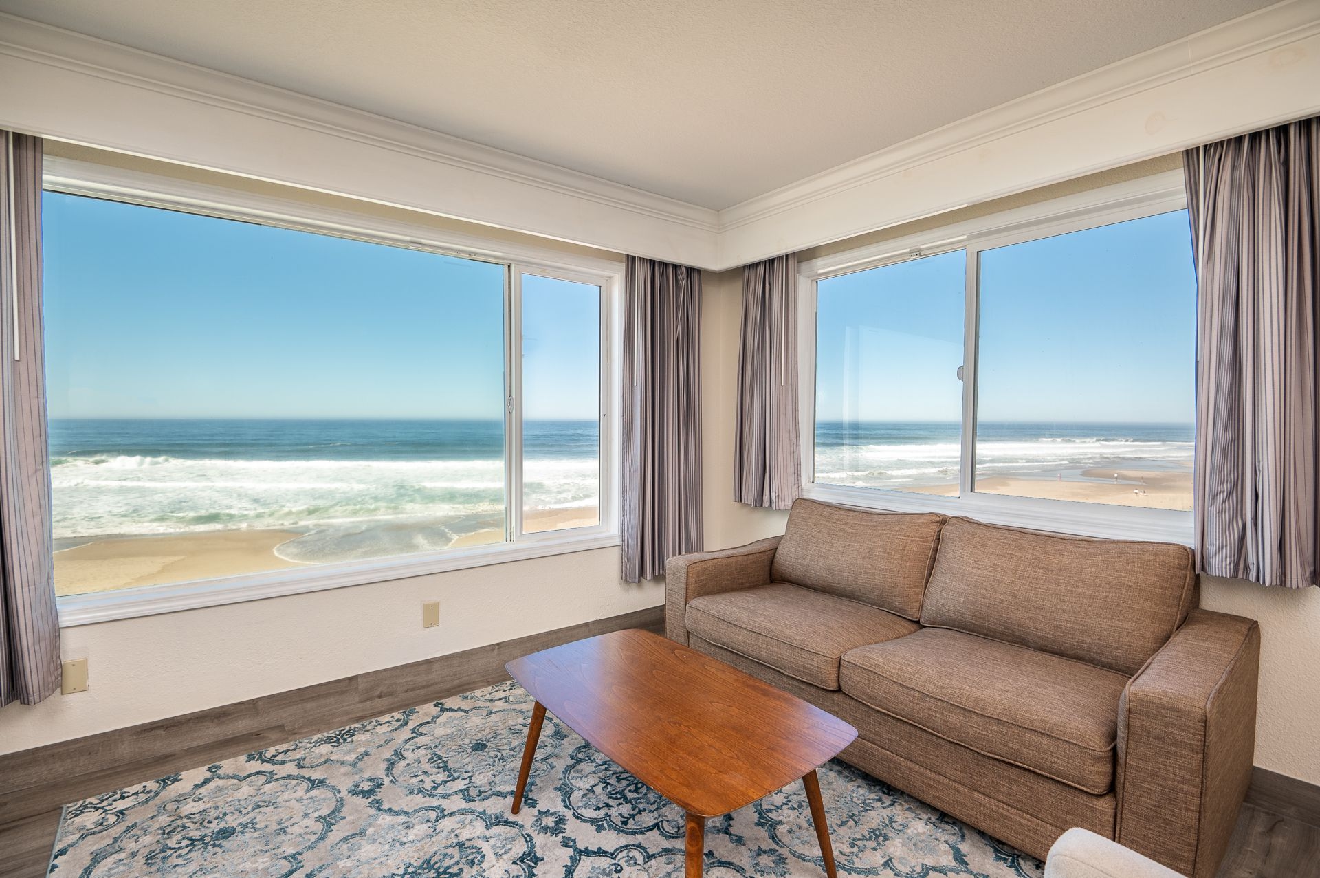 A brown couch and wooden coffee table on a patterned rug in a bright room with large windows overlooking the ocean.