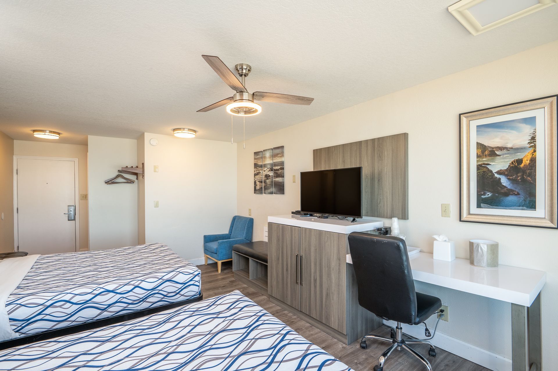 A hotel room with two beds, a television on a wooden cabinet, a desk with a chair, a blue armchair, and wall art.