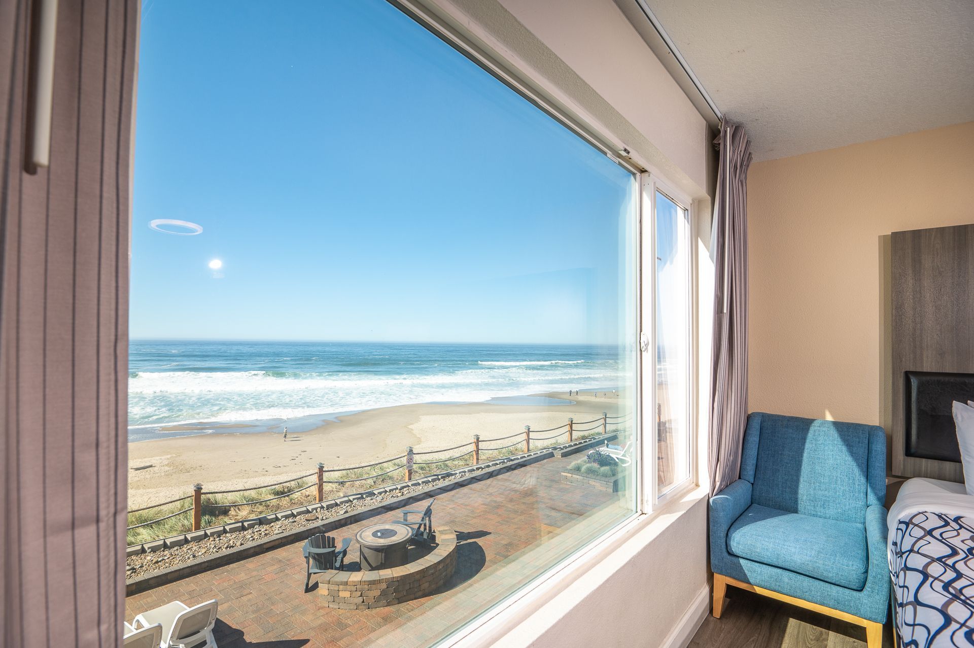 A room view through a large window overlooking a sunny beach, with a blue armchair in the foreground.