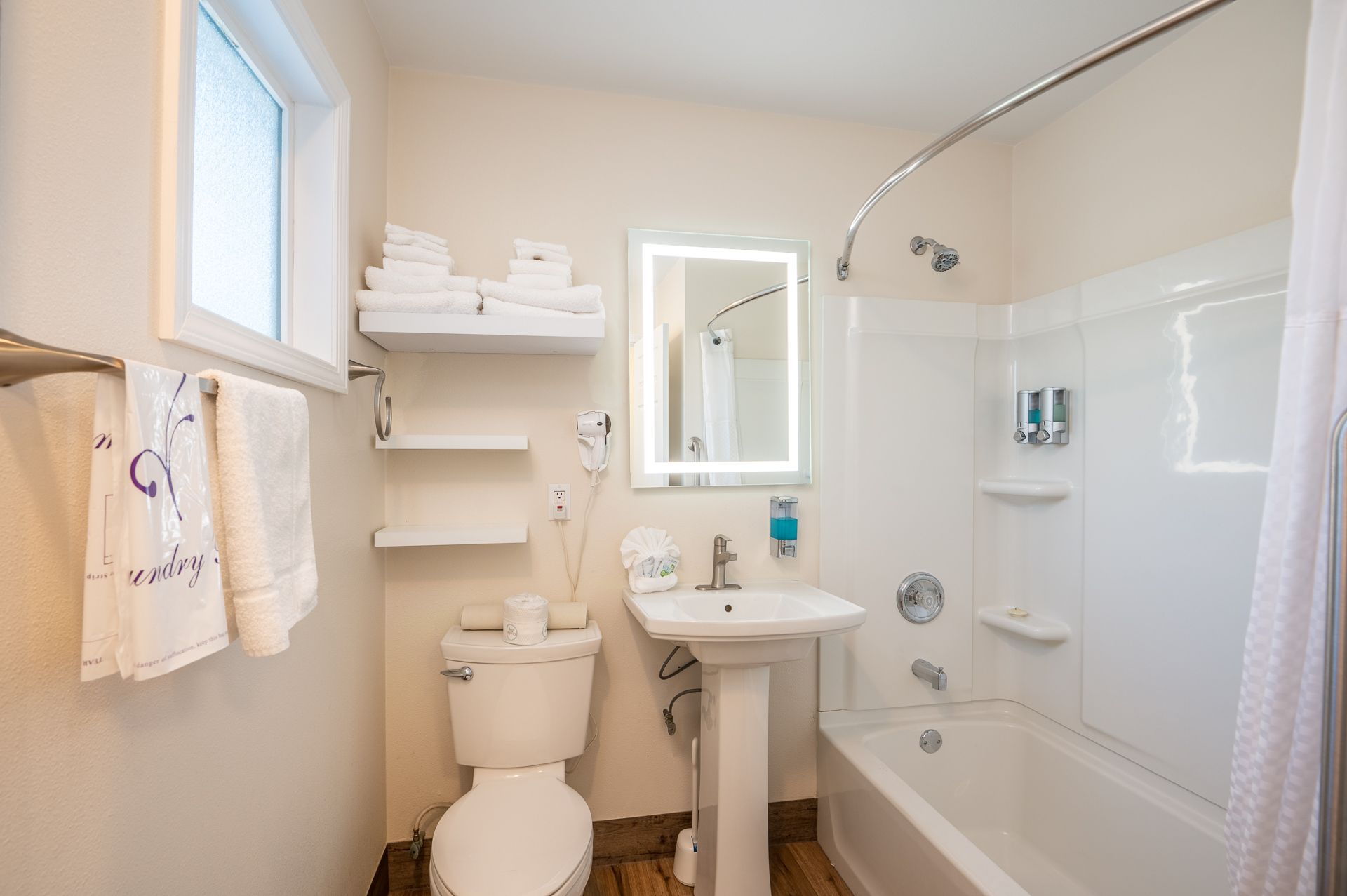 A neutral-toned hotel bathroom with a white pedestal sink, toilet, shower-tub combination, and open shelving with towels.