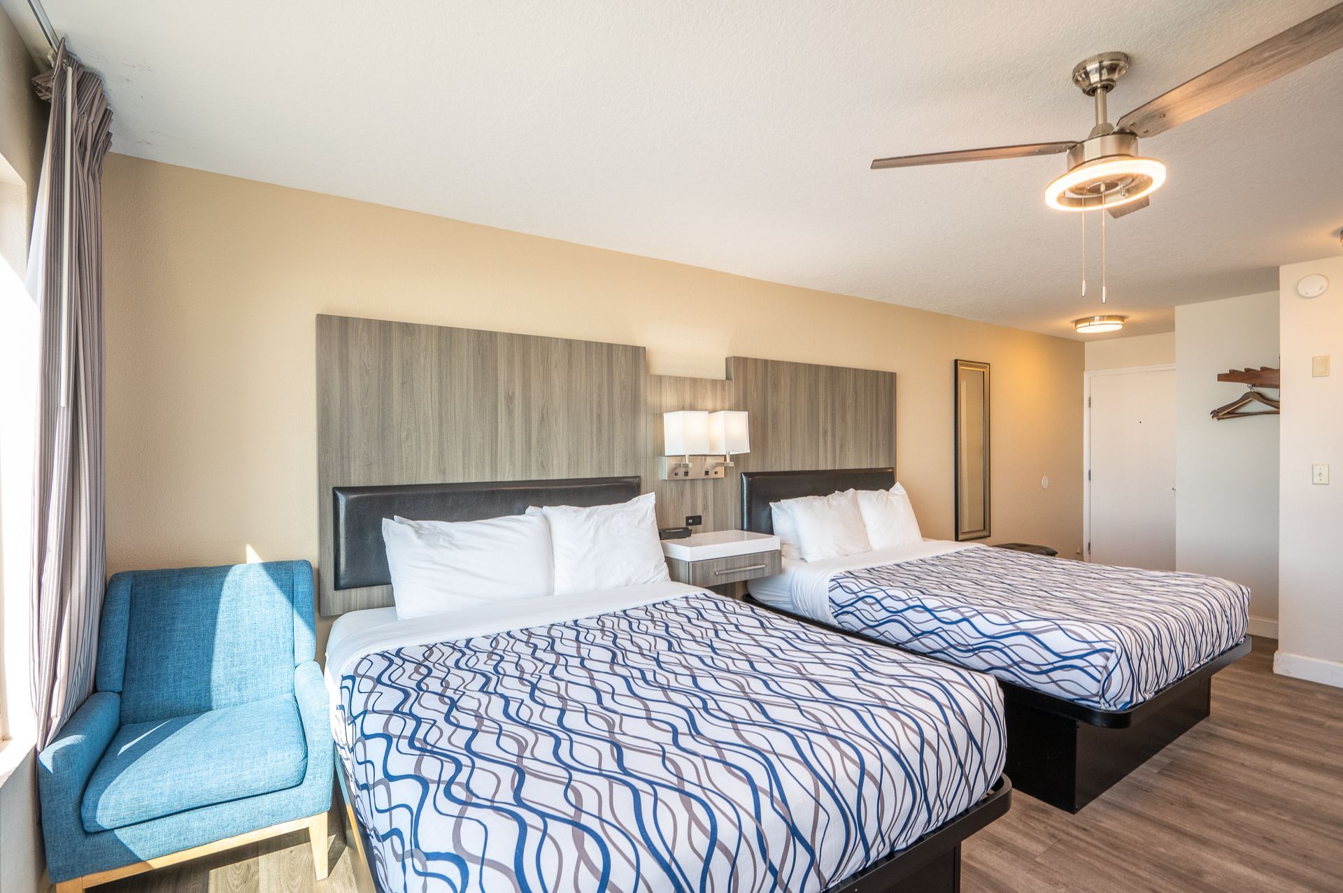 Hotel room with two queen beds featuring blue patterned comforters, a teal armchair, and a ceiling fan.