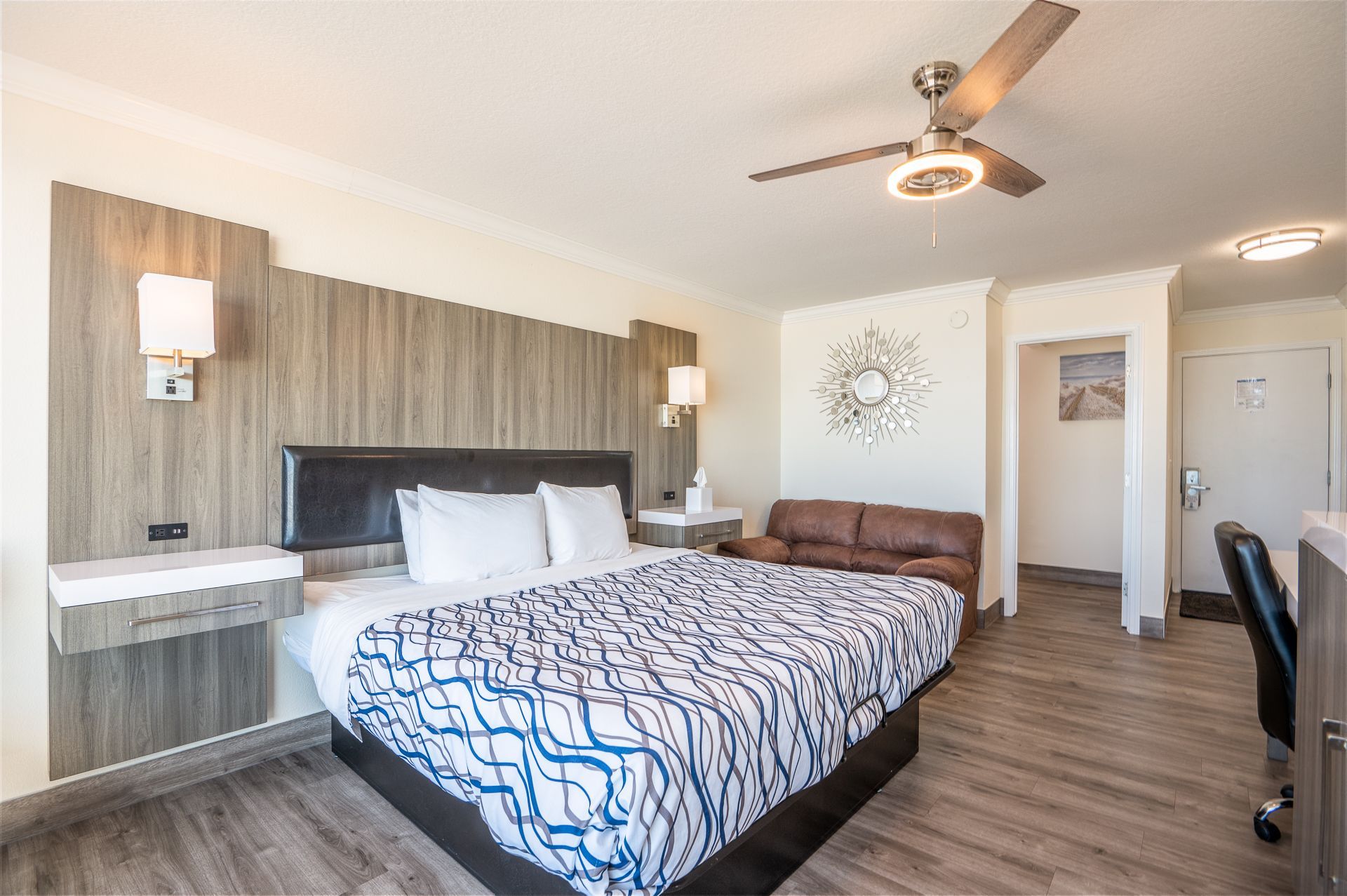 A hotel room featuring a king-size bed with a blue patterned duvet, a brown sofa, a ceiling fan, and light wood flooring.