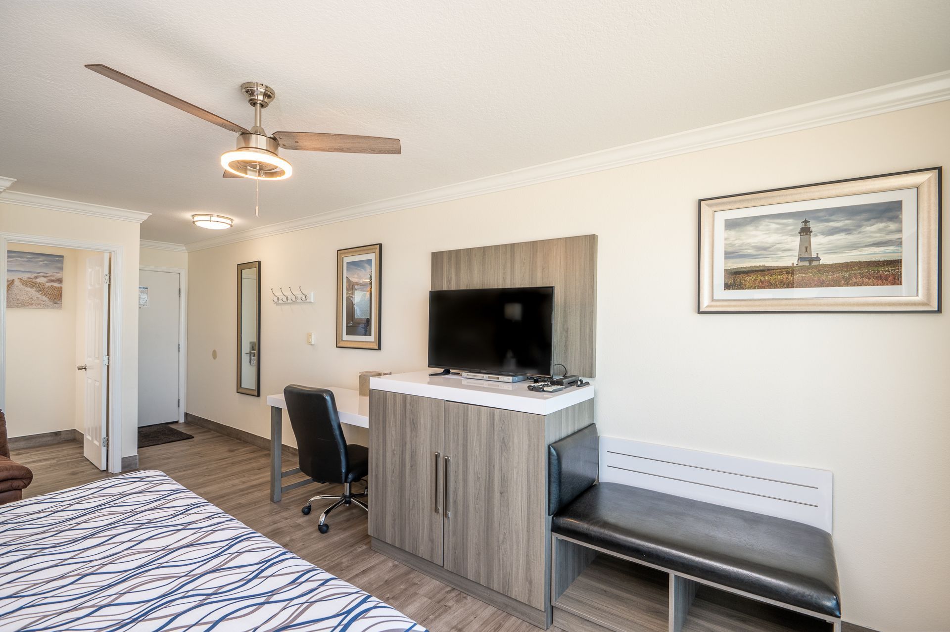 A hotel room with a bed, a desk with a black chair, a TV on a wooden console, and a black bench seat.