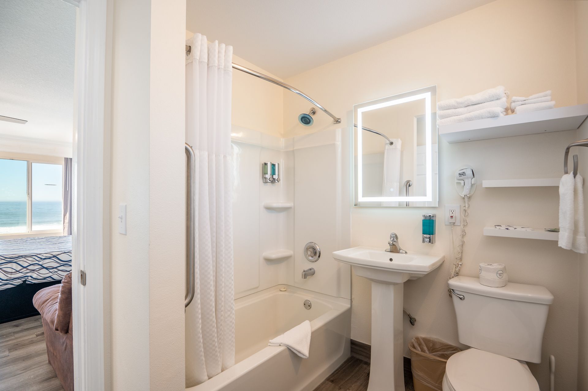 A clean hotel bathroom with a white pedestal sink, toilet, shower-tub combo, and illuminated mirror.