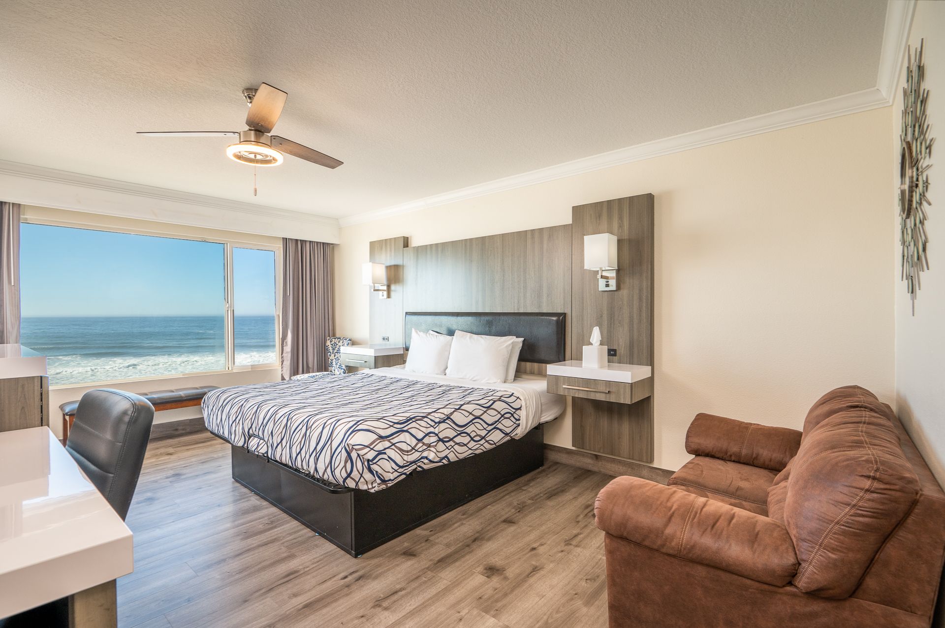 A hotel room with a queen bed, brown sofa, and a large window offering a scenic view of the ocean and beach.